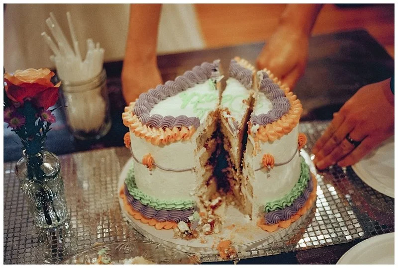 A partially sliced layered cake with orange, purple, and green frosting, on a silver cake board, with a bouquet of flowers and straws in the background.