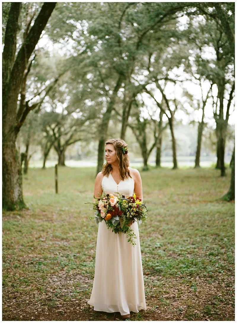 Bride standing in a wooded outdoor setting holding a large bouquet of flowers, wearing a white sleeveless wedding gown, with trees and greenery in the background.
