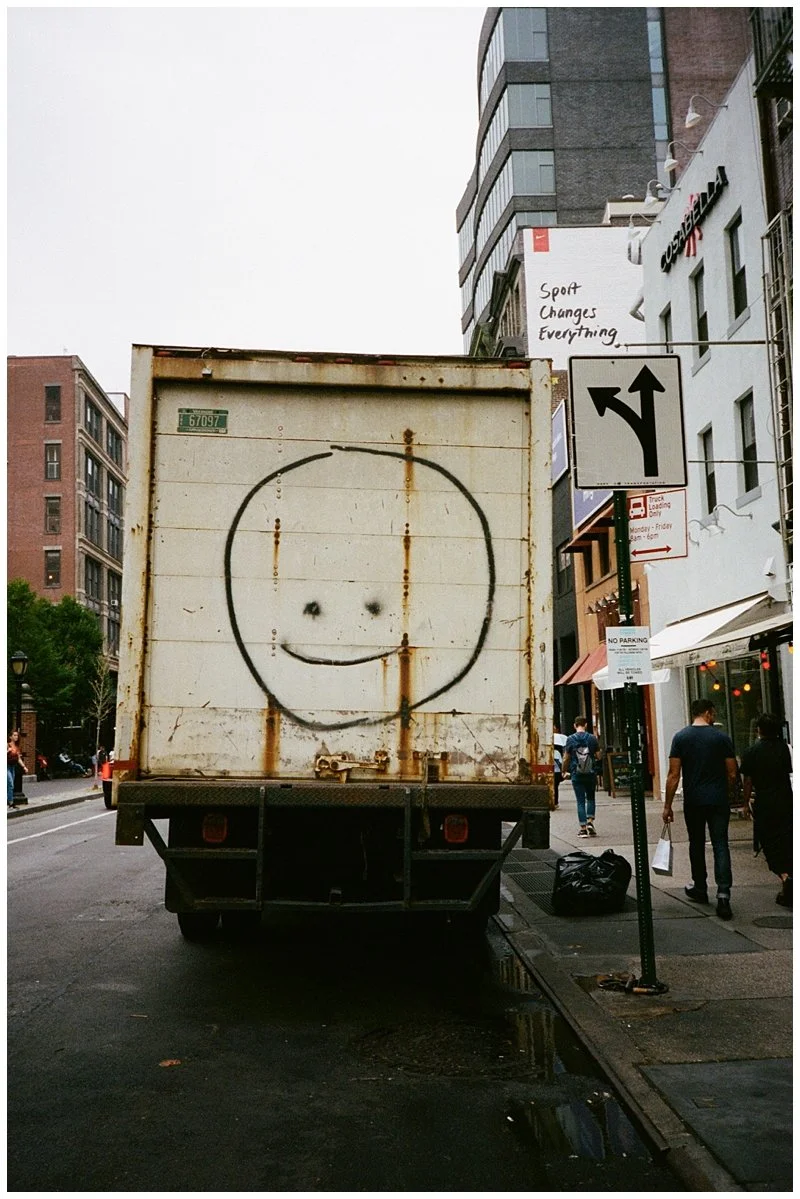 A rusty delivery truck with a simple smiley face drawn on the back, parked on a city street with pedestrians and tall buildings.