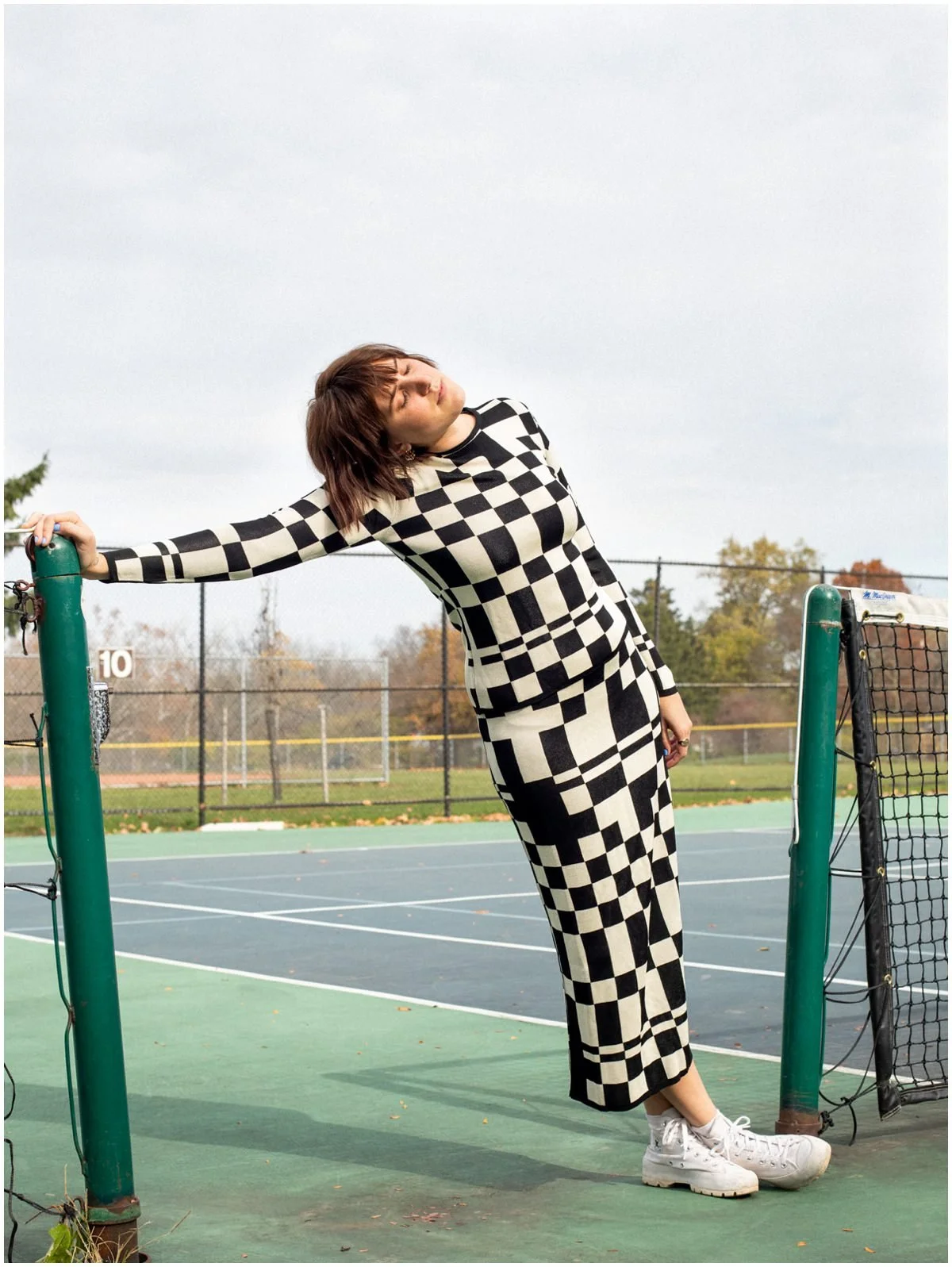A woman in a black and white geometric patterned outfit leaning on a green post at an outdoor tennis court