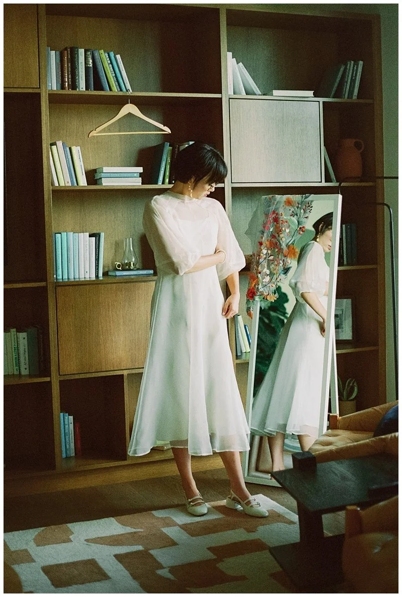 A woman in a white dress is standing in front of a large mirror in a room with wooden shelves filled with books, decorative items, and a hanging clothing hanger.
