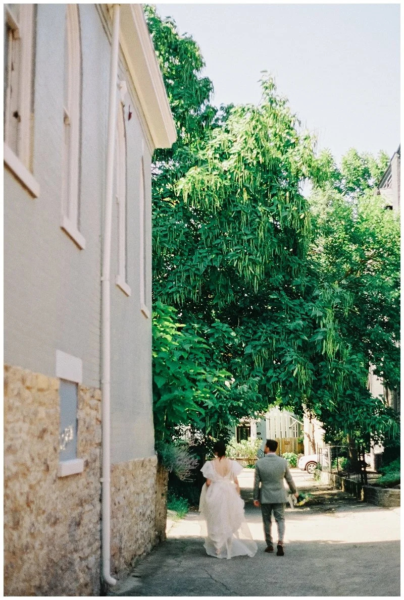 A bride and groom walking down a sidewalk in a residential neighborhood, with the bride wearing a white wedding gown and the groom in a gray suit, passing by a large green leafy tree and houses. Film Photography