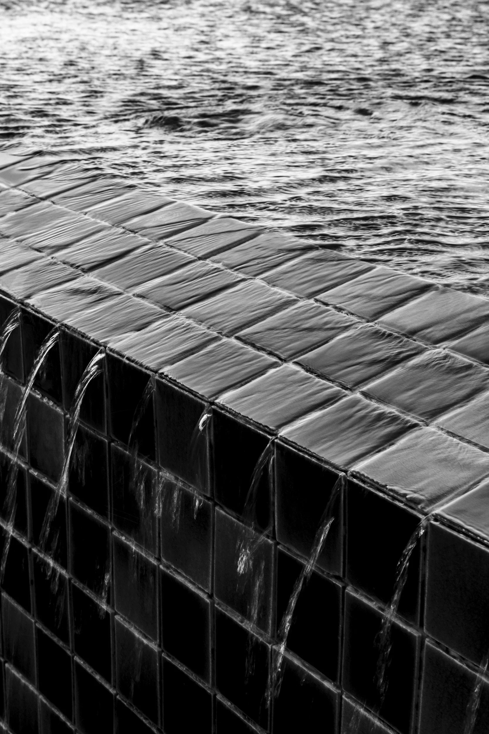 Black and white photo of a tiled water fountain with water flowing over the edge, overlooking a body of water.