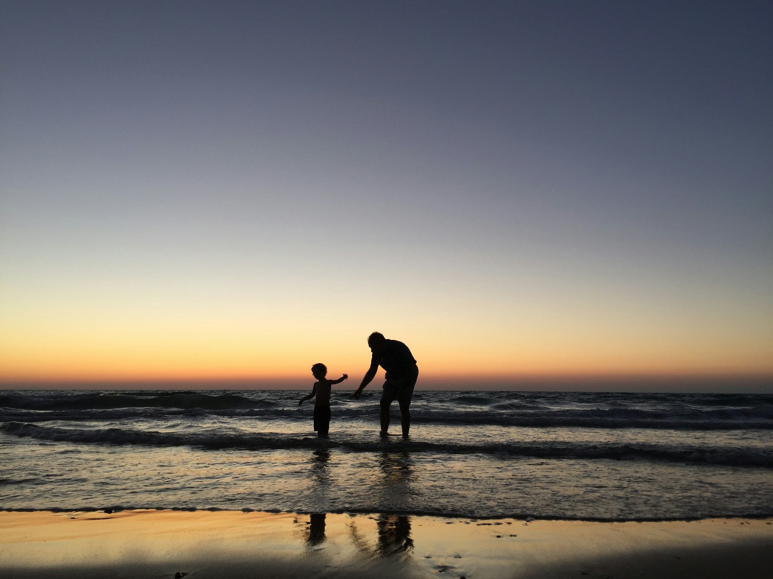 A silhouette of a person and a child holding hands in the shallow ocean water during sunset or sunrise.