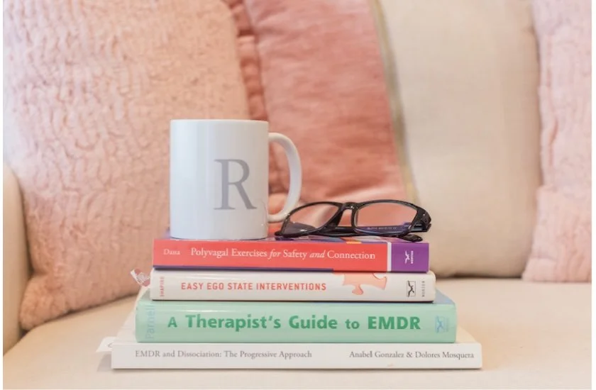 A stack of five books with a white mug and a pair of black-rimmed glasses on top, placed on a light-colored surface in front of pink and beige cushions.