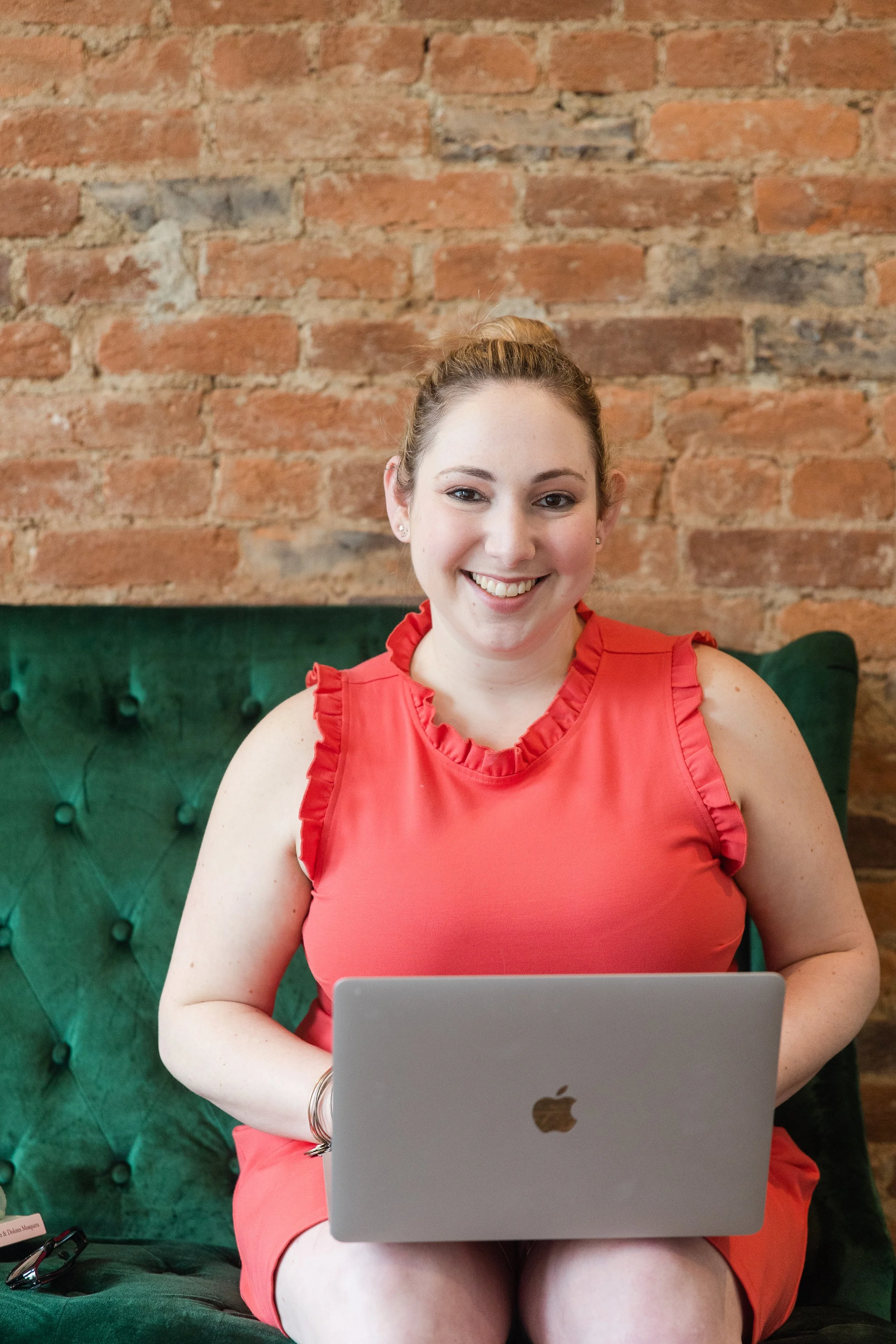 A woman with light brown hair in a bun, wearing a red sleeveless ruffled top, sitting on a green tufted chair, smiling, holding a silver MacBook in front of her, with a brick wall background.