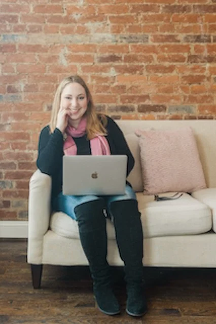 A woman sitting on a cream sofa with a laptop on her lap, in front of a brick wall, smiling, wearing a pink scarf, black sweater, and black boots.
