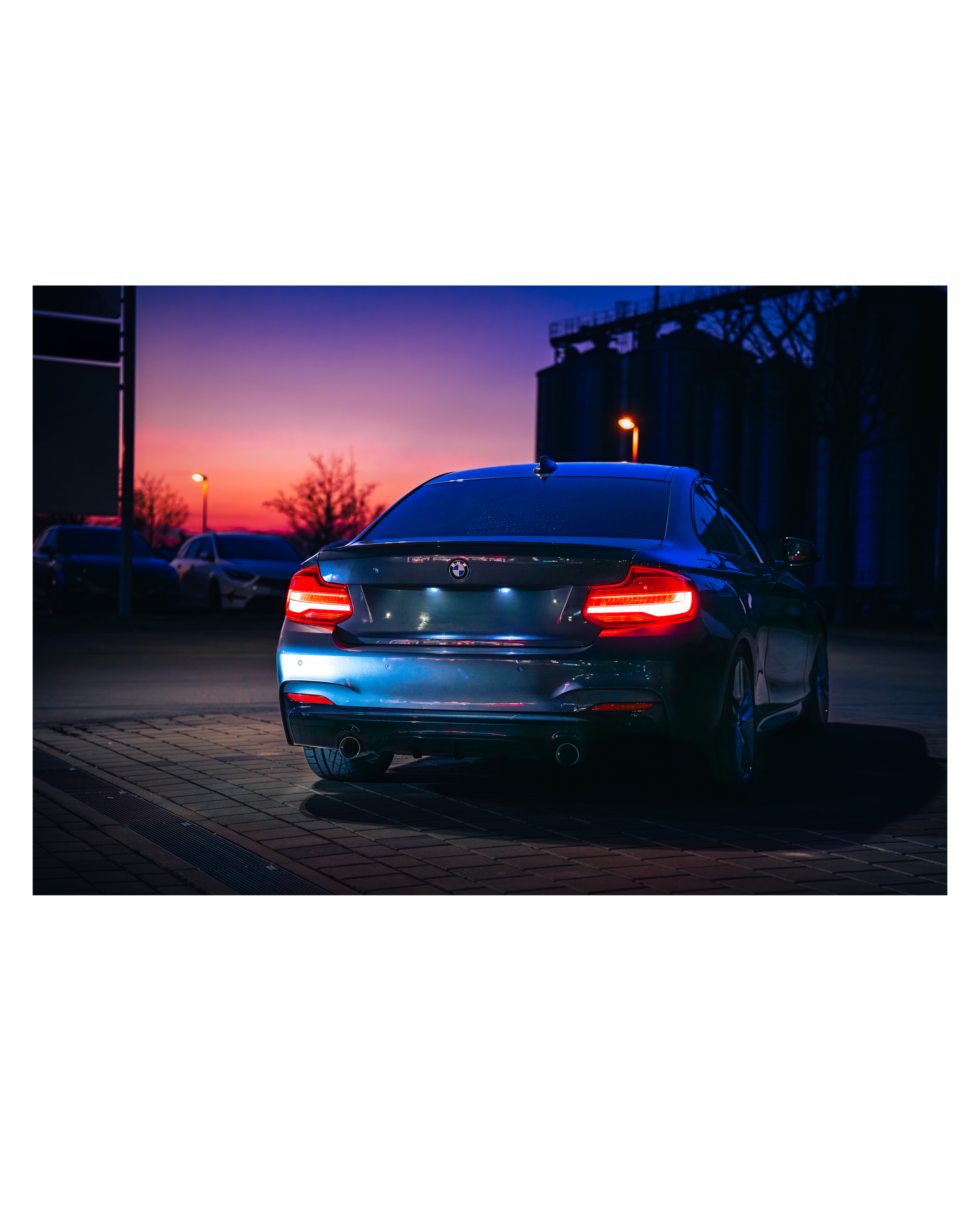 Ein dunkler BMW vor einem Sonnenuntergang mit rosa, lila und blauen Himmel, geparkte Fahrzeuge im Hintergrund und eine Industrieanlage rechts