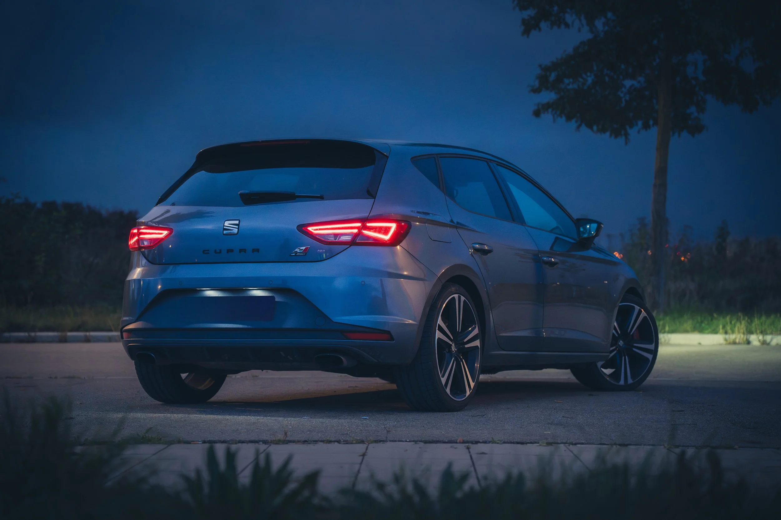 Ein blauer Seat Cupra-Auto wird nachts geparkt, mit leuchtenden Rücklichtern und einem Baum im Hintergrund.