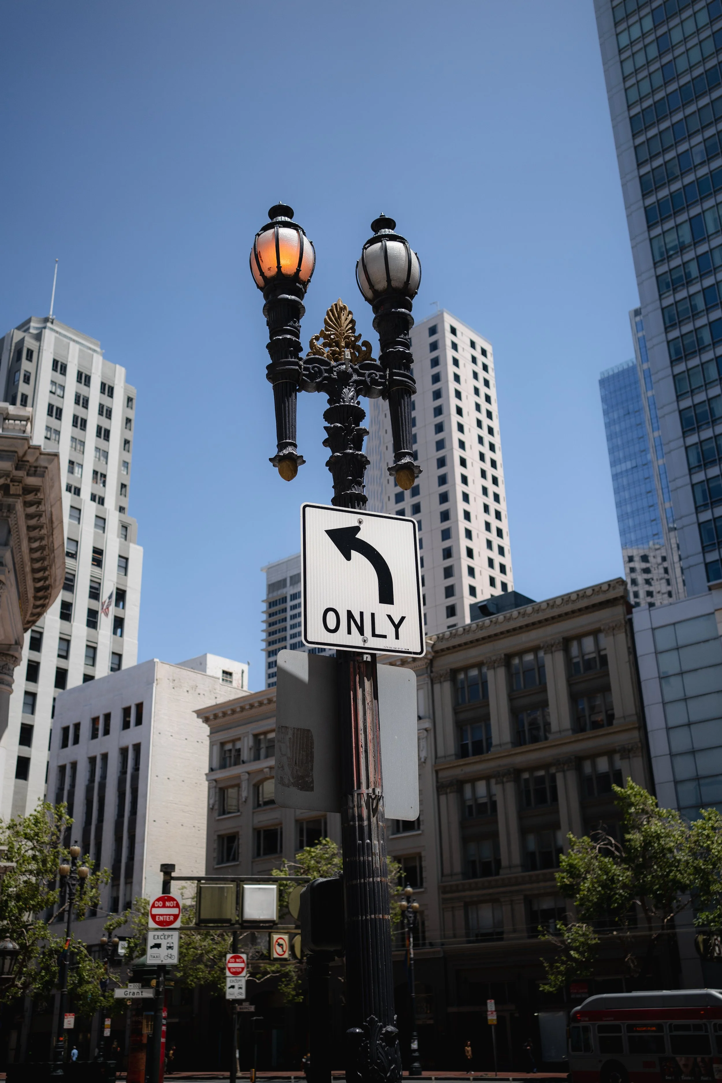 Straßenlaterne mit einem Verkehrszeichen, das nach links abbiegt, und Hochhäuser in einer Stadt bei sonnigem Wetter.