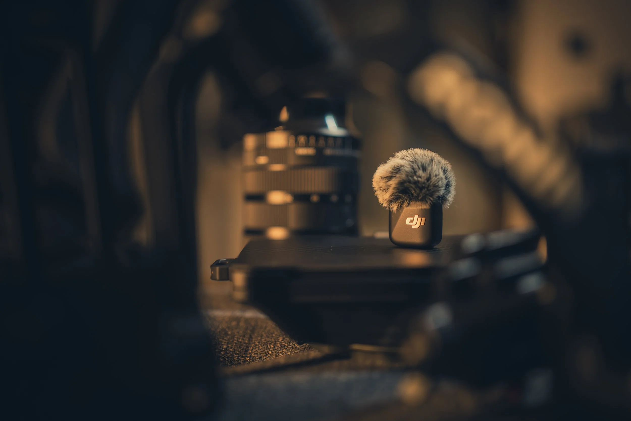 Mikrofon mit Objektiv auf einer schwarzen Oberfläche, im Hintergrund technische Ausrüstung, warme Beleuchtung.