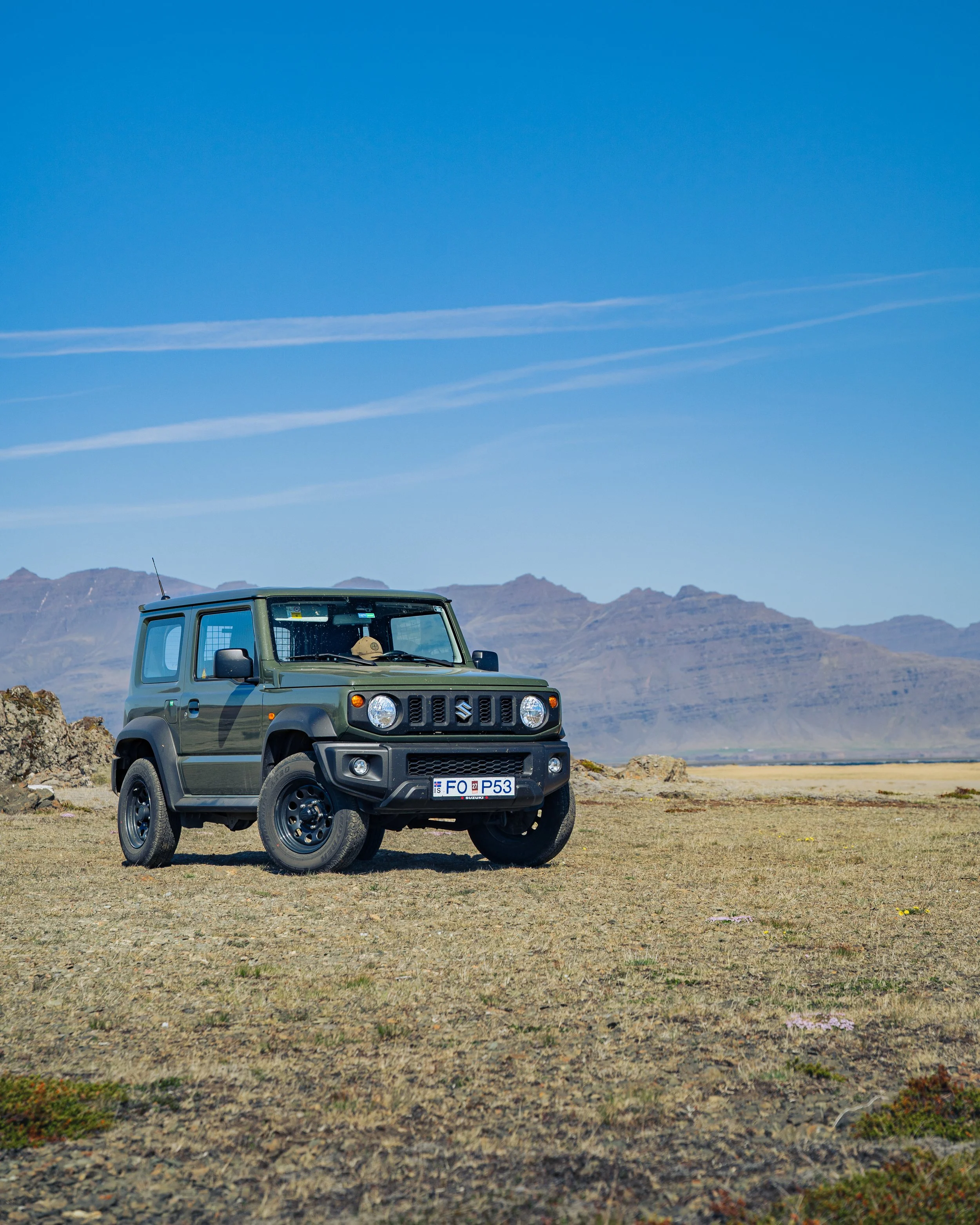 Ein grüner Suzuki Jimny steht auf einer offenen, kargen Landschaft mit Bergen im Hintergrund und einem klaren blauen Himmel darüber.
