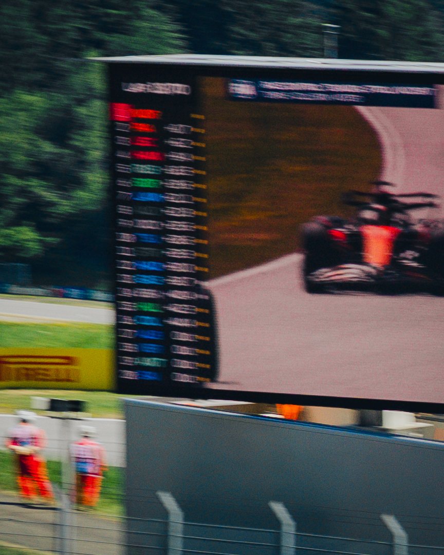 Ein großer Fernseher zeigt ein Rennsportszene mit einem roten Formelrennwagen auf einer Rennstrecke. Im Hintergrund sind Bäume und jemand in roten Kleidung, vermutlich Streckenposten, zu sehen.