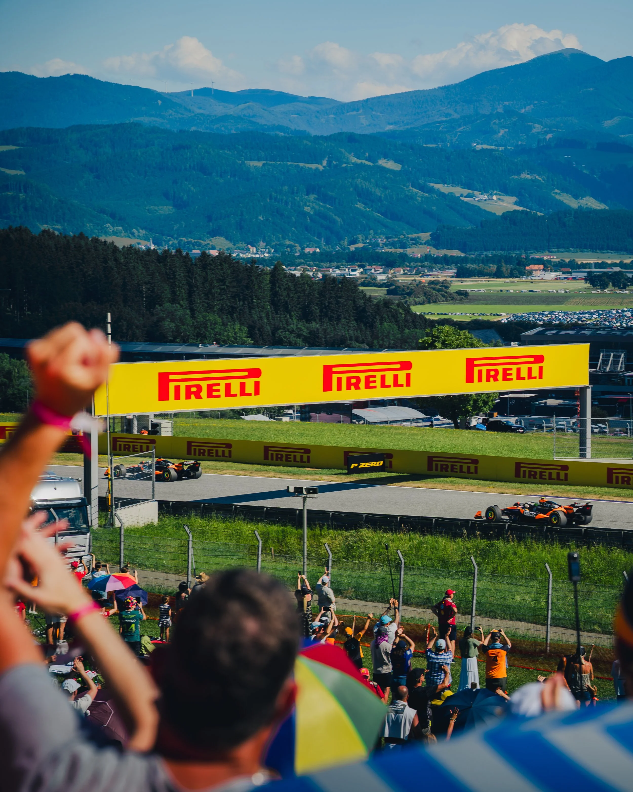 Motorsportrennen mit Flucht von Formelwagen auf einer Rennstrecke im Grünen, Zuschauer beobachten von der Seite, Berge im Hintergrund, bewölkter Himmel.