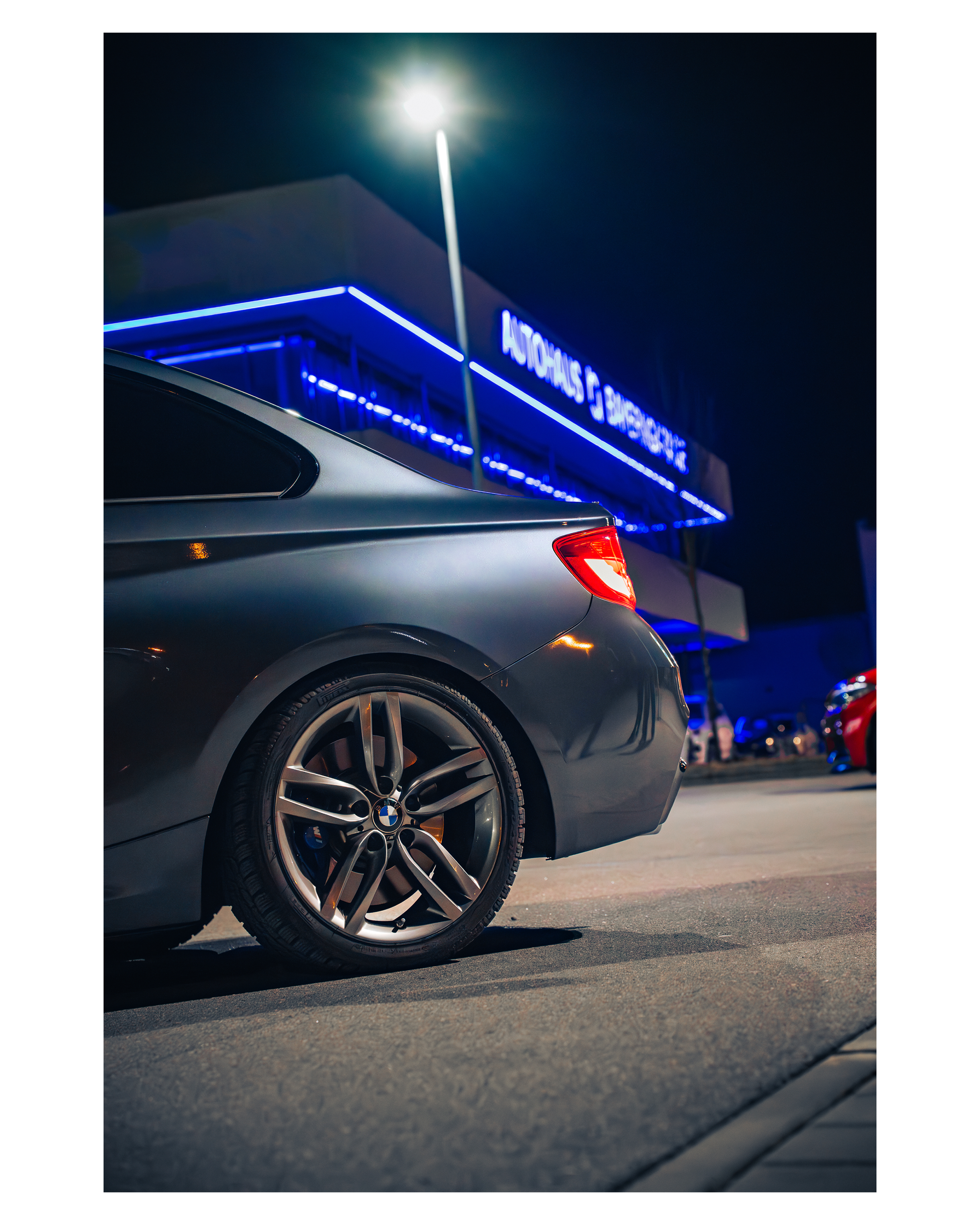 Nachtaufnahme eines grauen BMW-Autos vor einem blauen Neon-Laden mit der Beschriftung "Autohaus & Service" in einem Autohof.