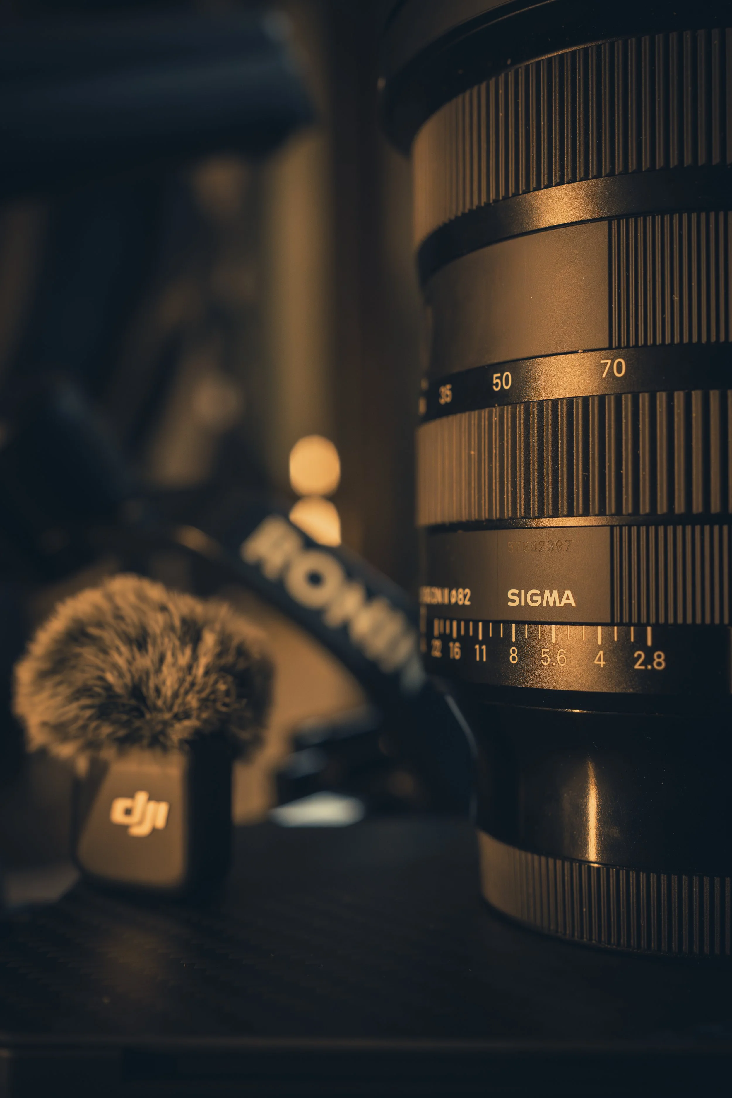 Nahaufnahme eines Sigma Kameraobjektivs, mit einer Mikrofon und einer Kamera-Anti-Schüttelmaske auf einem dunklen Oberflächen in warmem Licht.