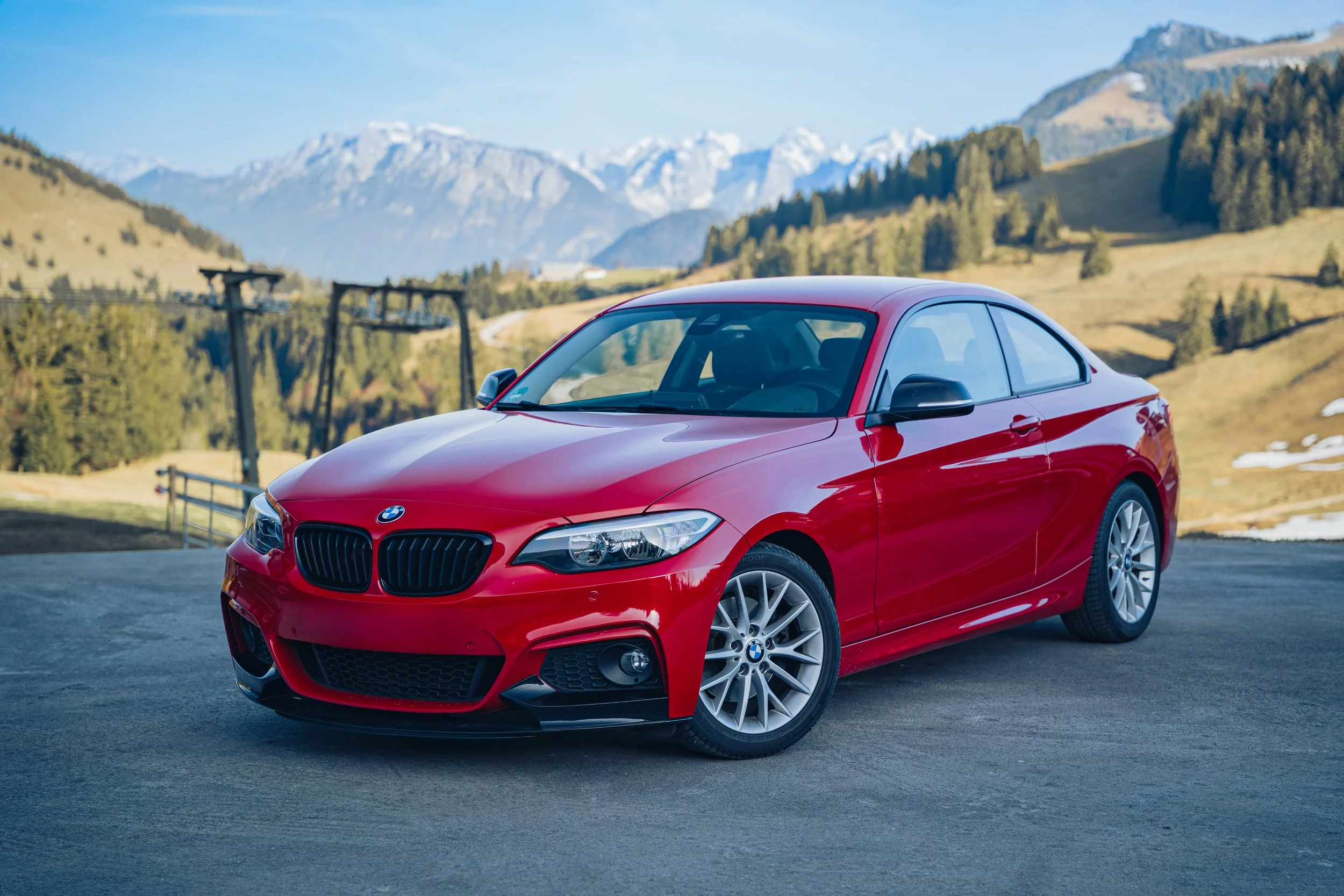 Roter BMW-Cabrio auf asphaltierter Straße vor malerischer Berglandschaft mit Schnee bedeckten Gipfeln und grünen Hügeln.