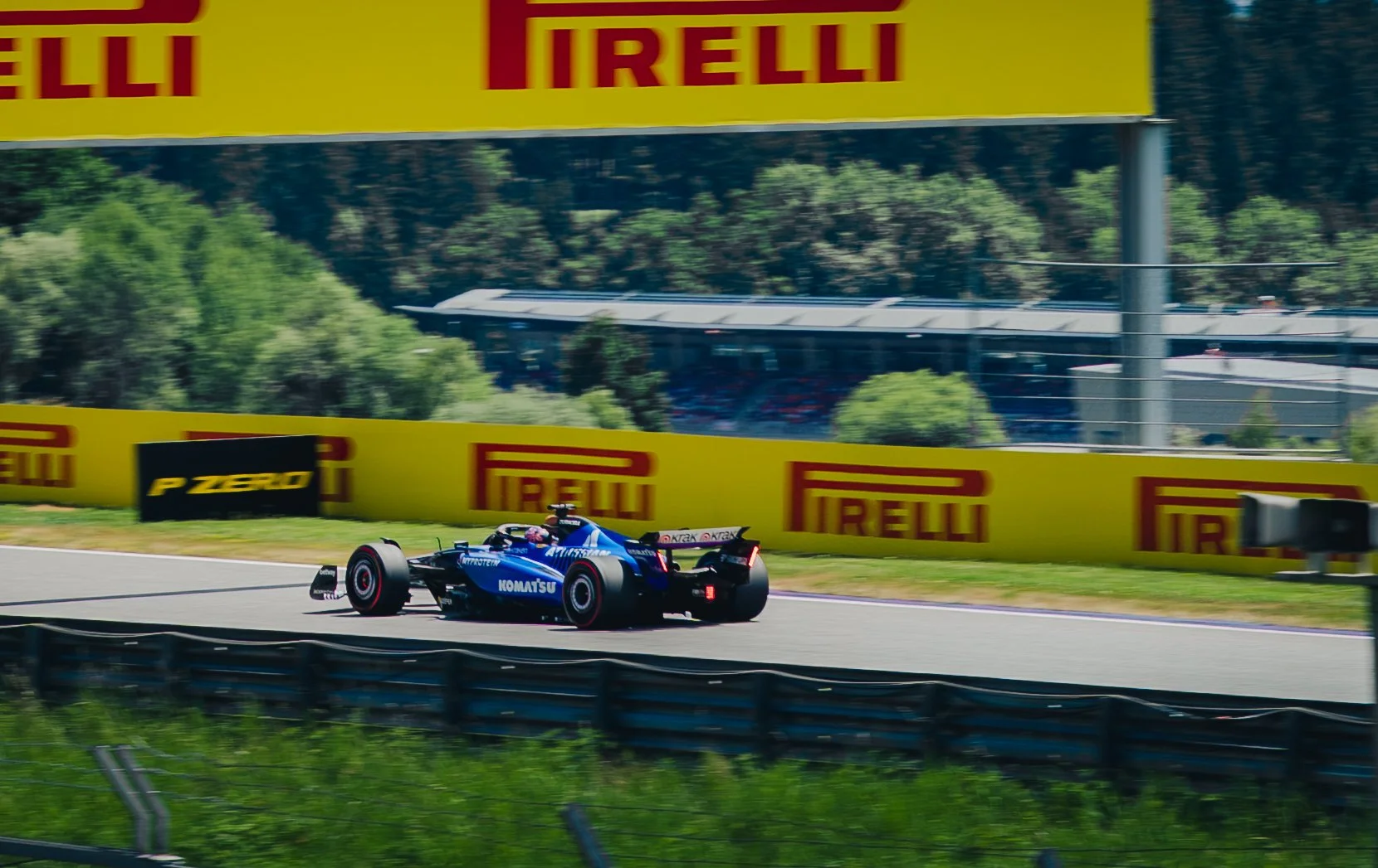 Ein Formel-1-Rennauto auf einer Rennstrecke, umgeben von Werbetafeln mit Pirelli und P Zero Anzeigen, während Bäume im Hintergrund zu sehen sind.