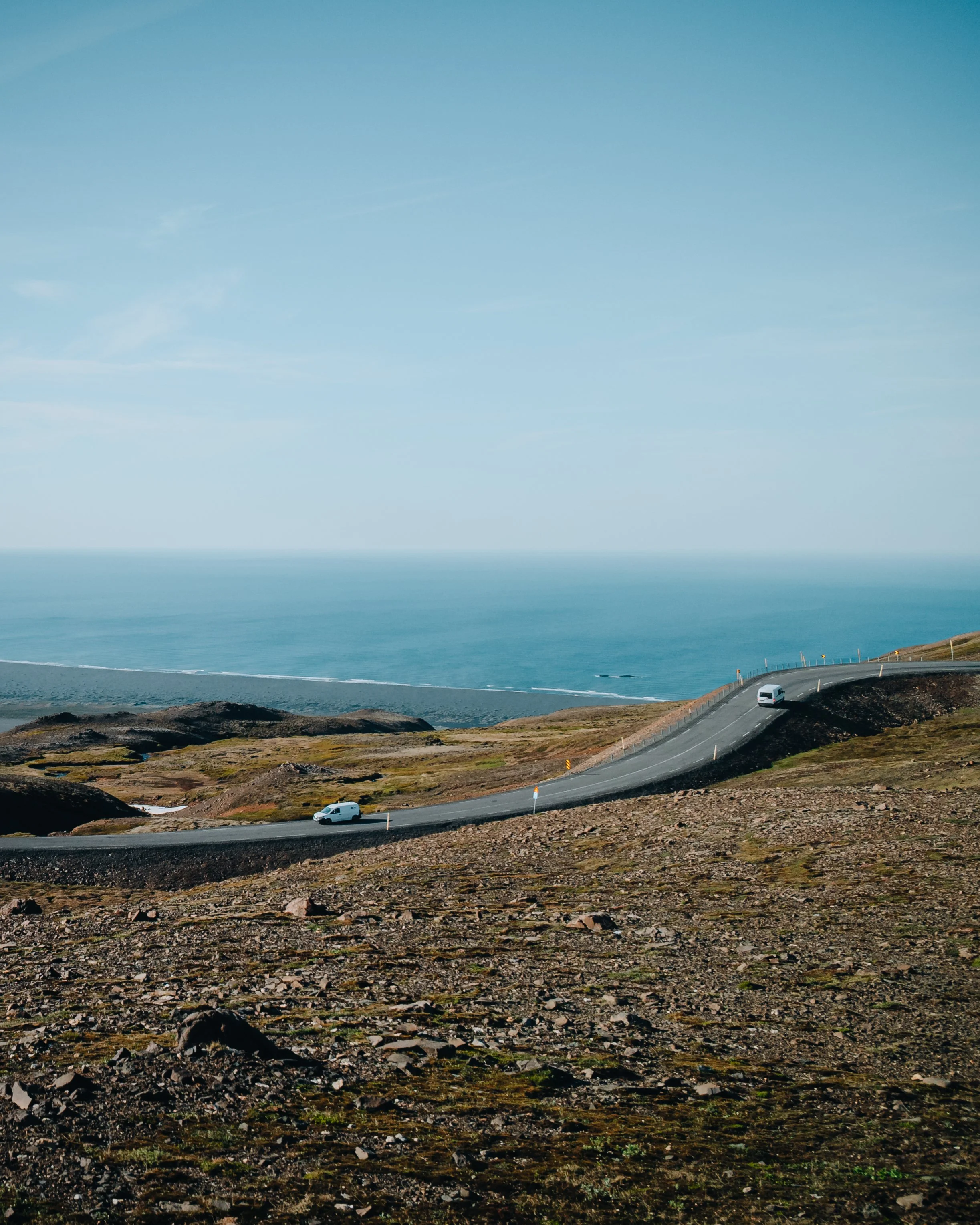 Straße an der Küste mit drei weißen Autos, umgeben von felsigem, grünlichem Gelände und Blick auf den Ozean im Hintergrund unter blauem Himmel.