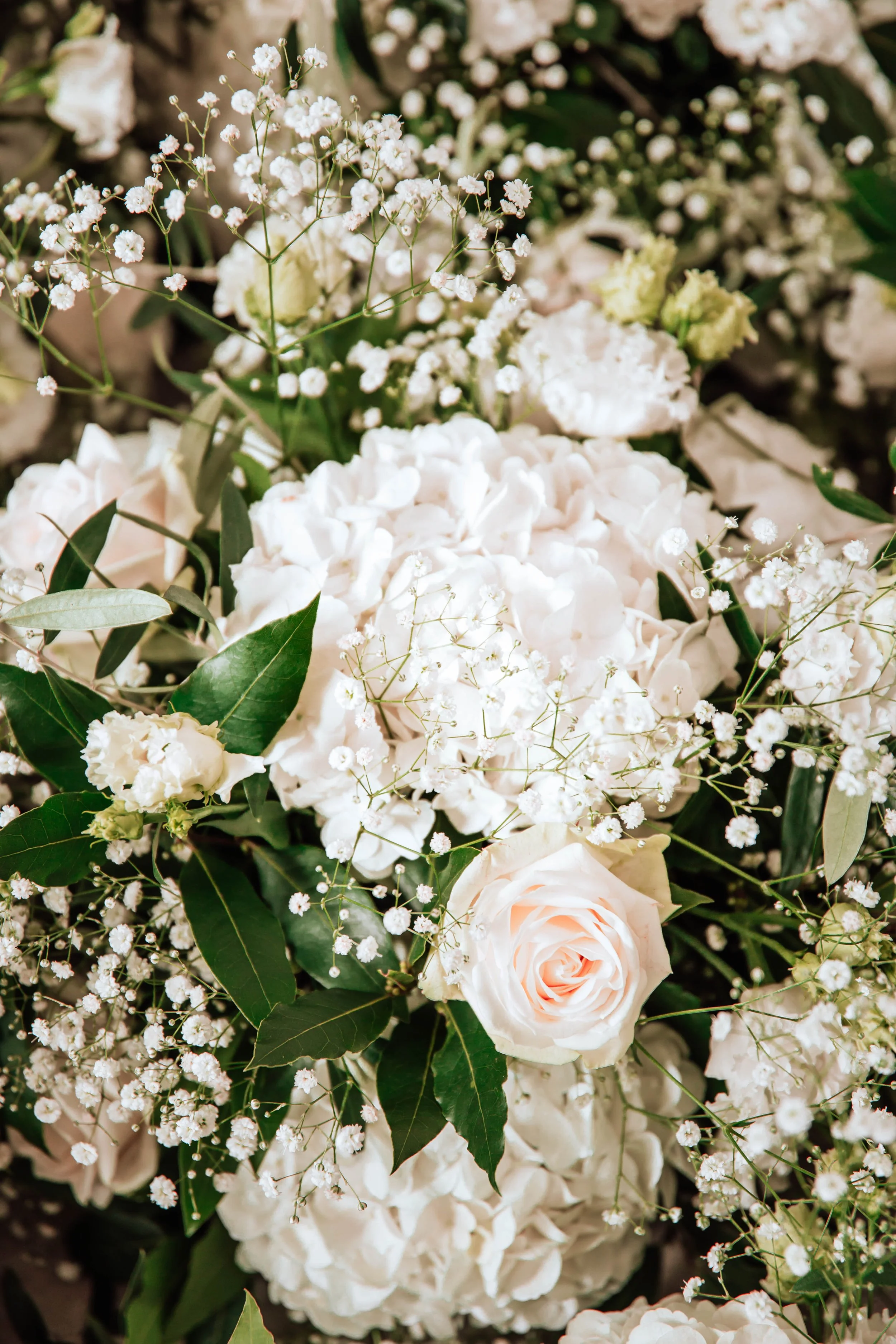 Bouquet de fleurs blanches comprenant des roses, des hortensias, et des petites fleurs de gypsophile avec des feuilles vertes.