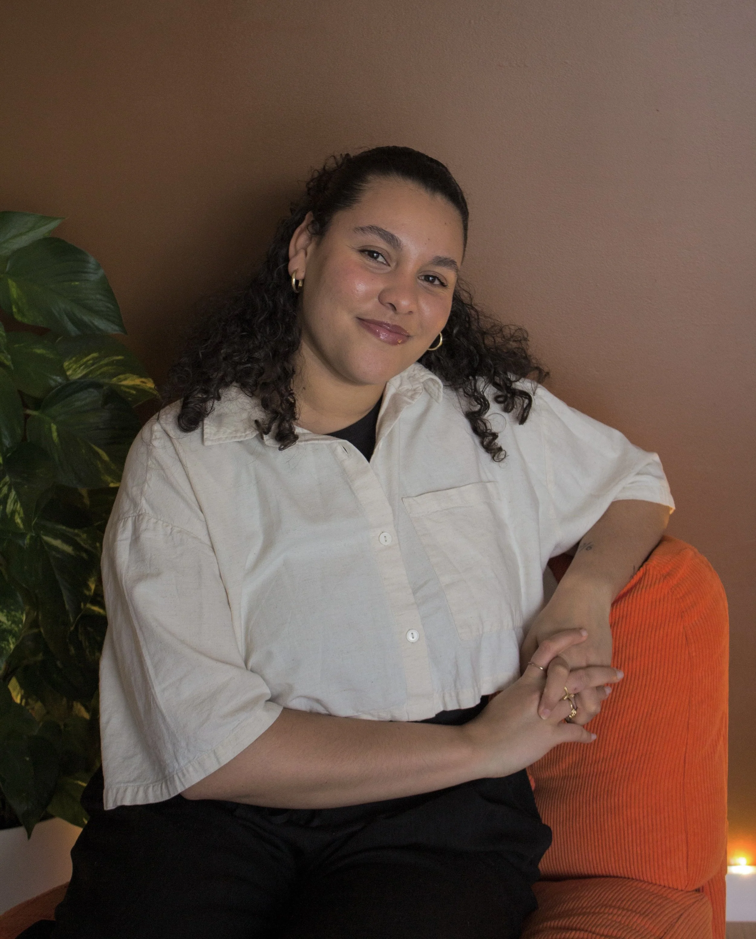 Une femme souriante assise sur un canapé orange, portant une chemise beige, avec un mur marron en arrière-plan.
