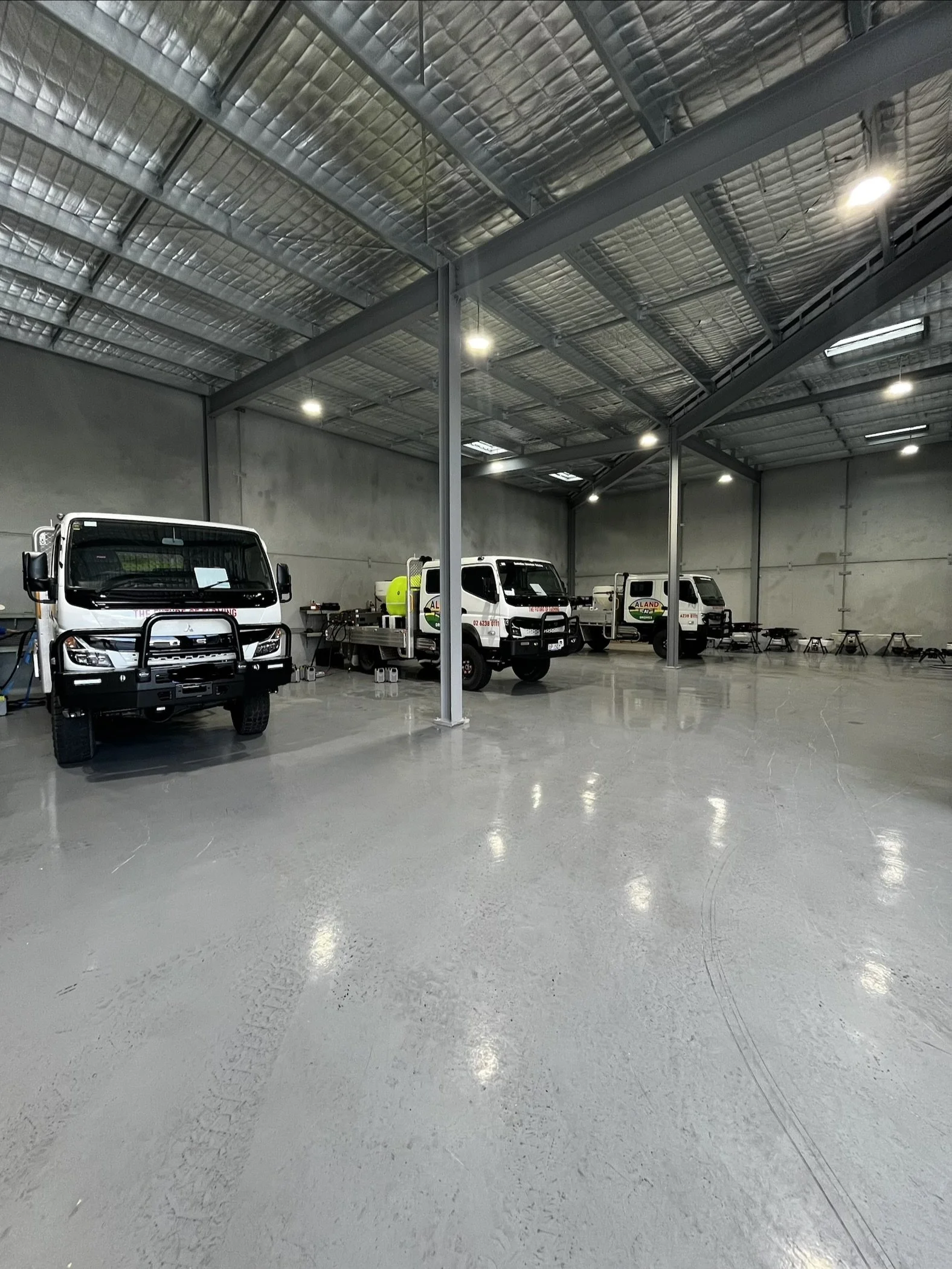 Interior view of a large warehouse or garage with three white trucks parked inside, some chairs and tables against the walls, and bright overhead lighting.
