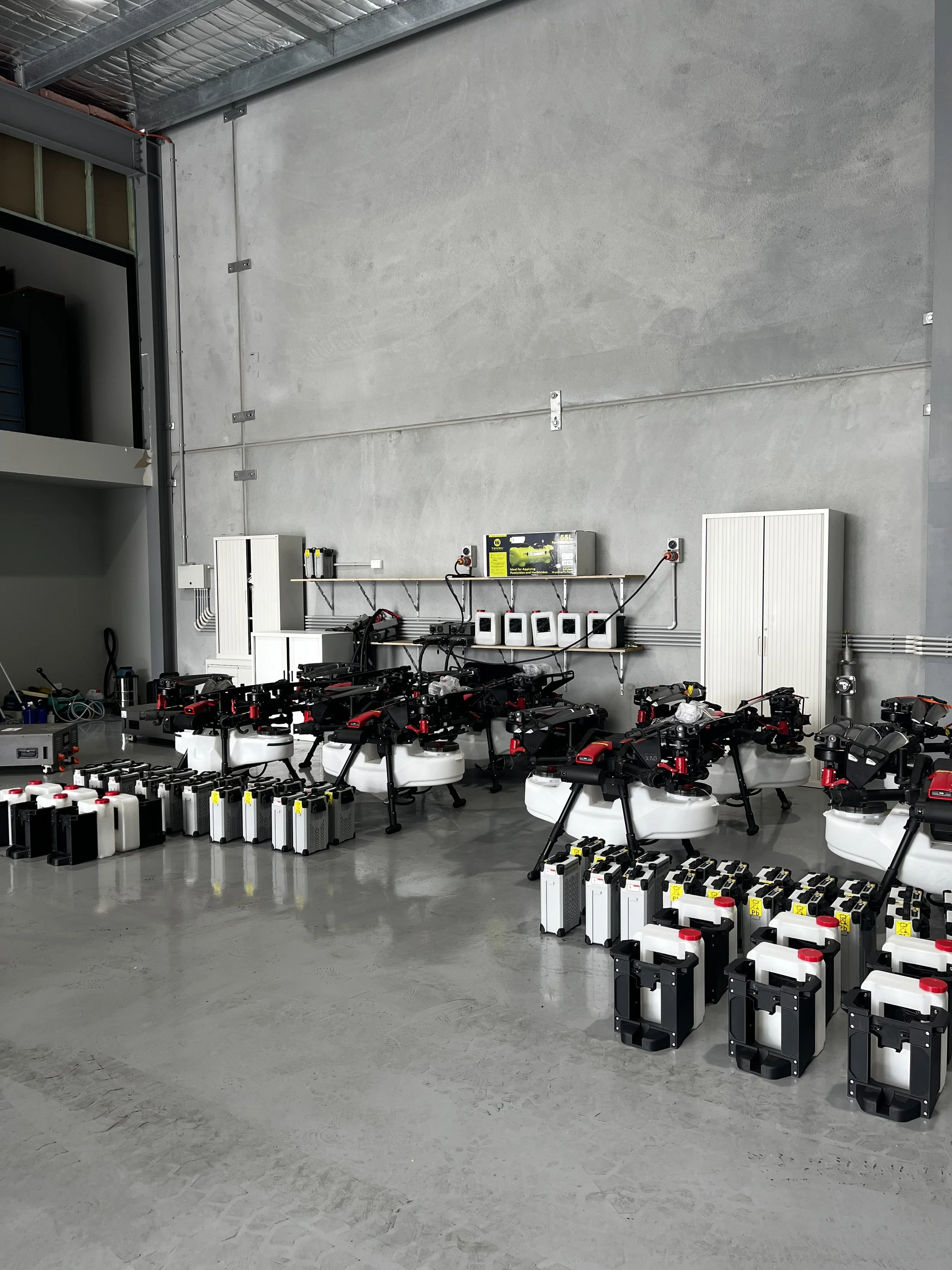 Collection of drones and drone batteries in a warehouse or workshop with a concrete floor and gray wall.