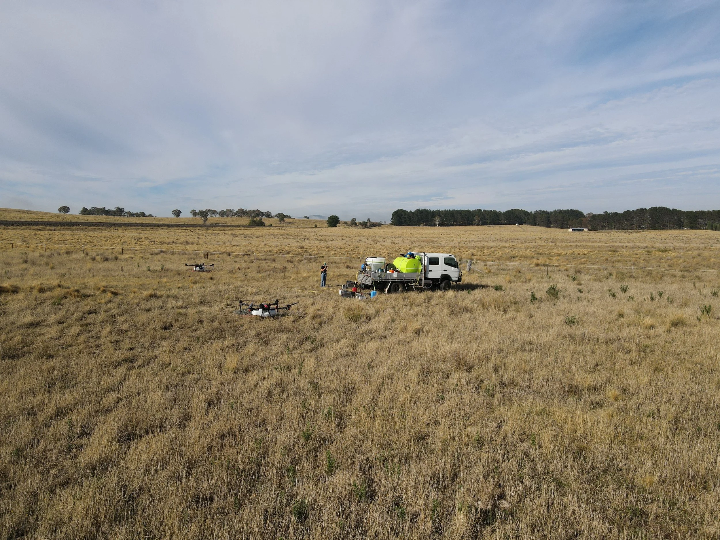 A drone operator in a field with a drone on the ground and a truck carrying equipment, under a partly cloudy sky.