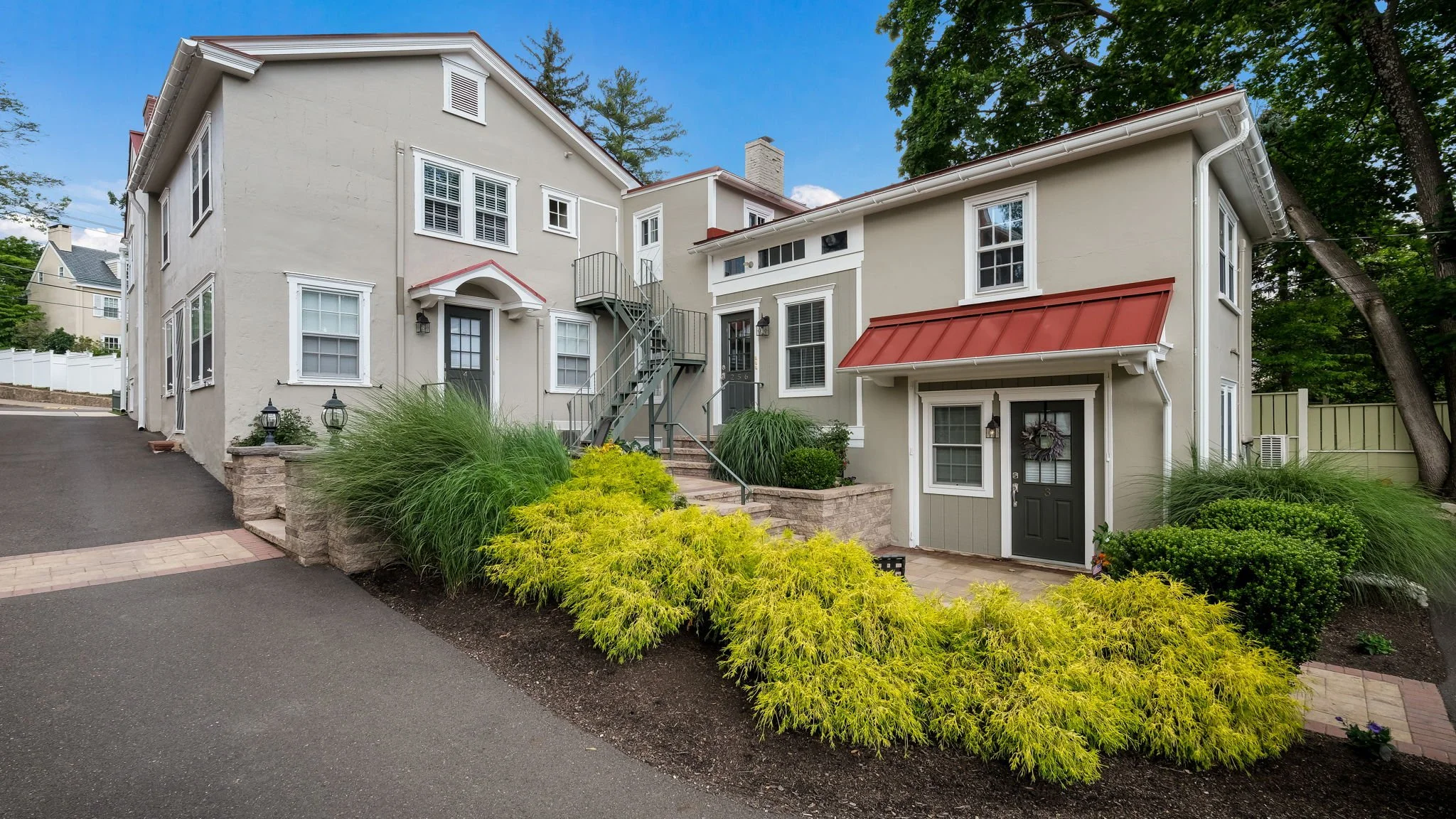 Exterior view of a beige residential building with multiple windows, a staircase, and landscaped front yard with yellow and green bushes.