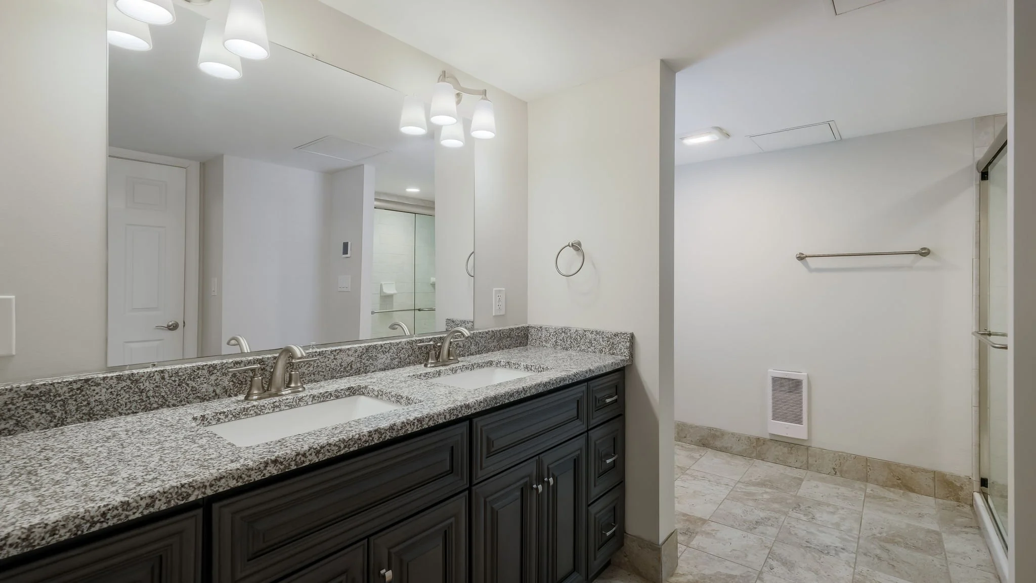 Bathroom with a granite countertop double vanity, a large mirror, and a wall-mounted light fixture. The space includes a walk-in shower with glass door, towel bars, and a vent on the wall. The floor is tiled in beige, and the walls are painted white.