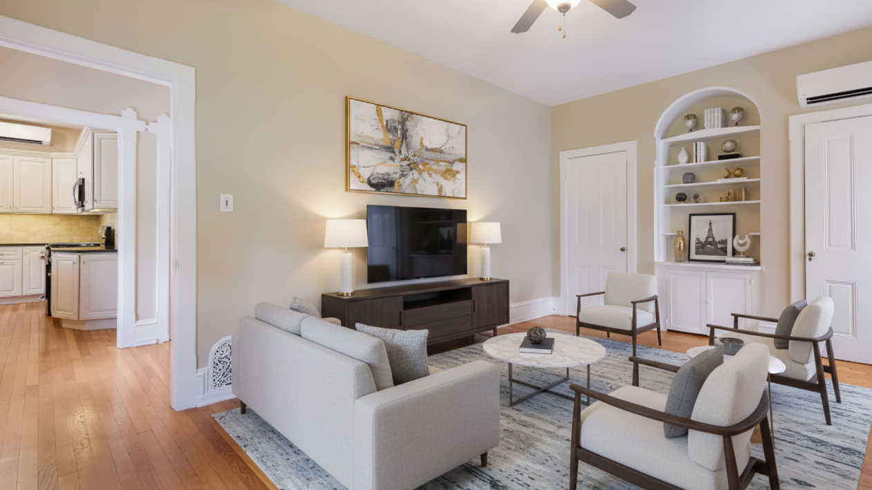 Living room with beige walls, a white ceiling fan, a wall-mounted TV above a wooden console, and a light-colored sofa and armchairs arranged around a circular marble coffee table. There are decorative shelves with various items and a doorway leading to a kitchen.