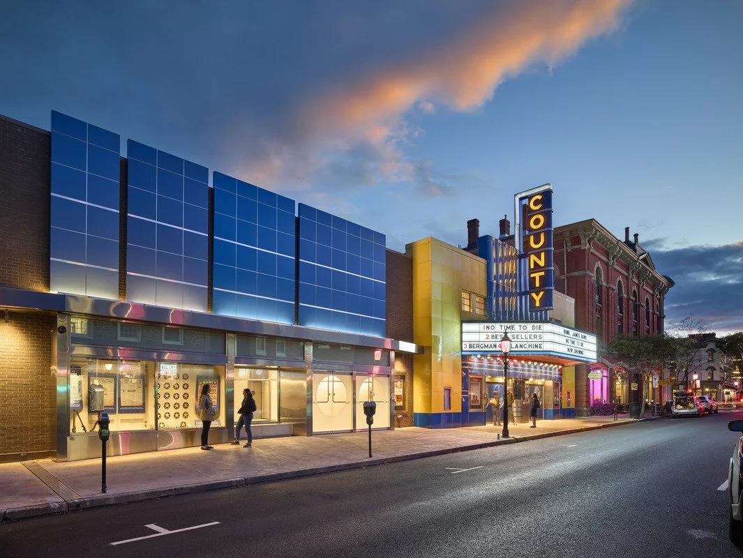 Night view of a small-town movie theater with a marquee and illuminated 'COUNTY' sign, people standing outside, and a colorful sky with clouds.
