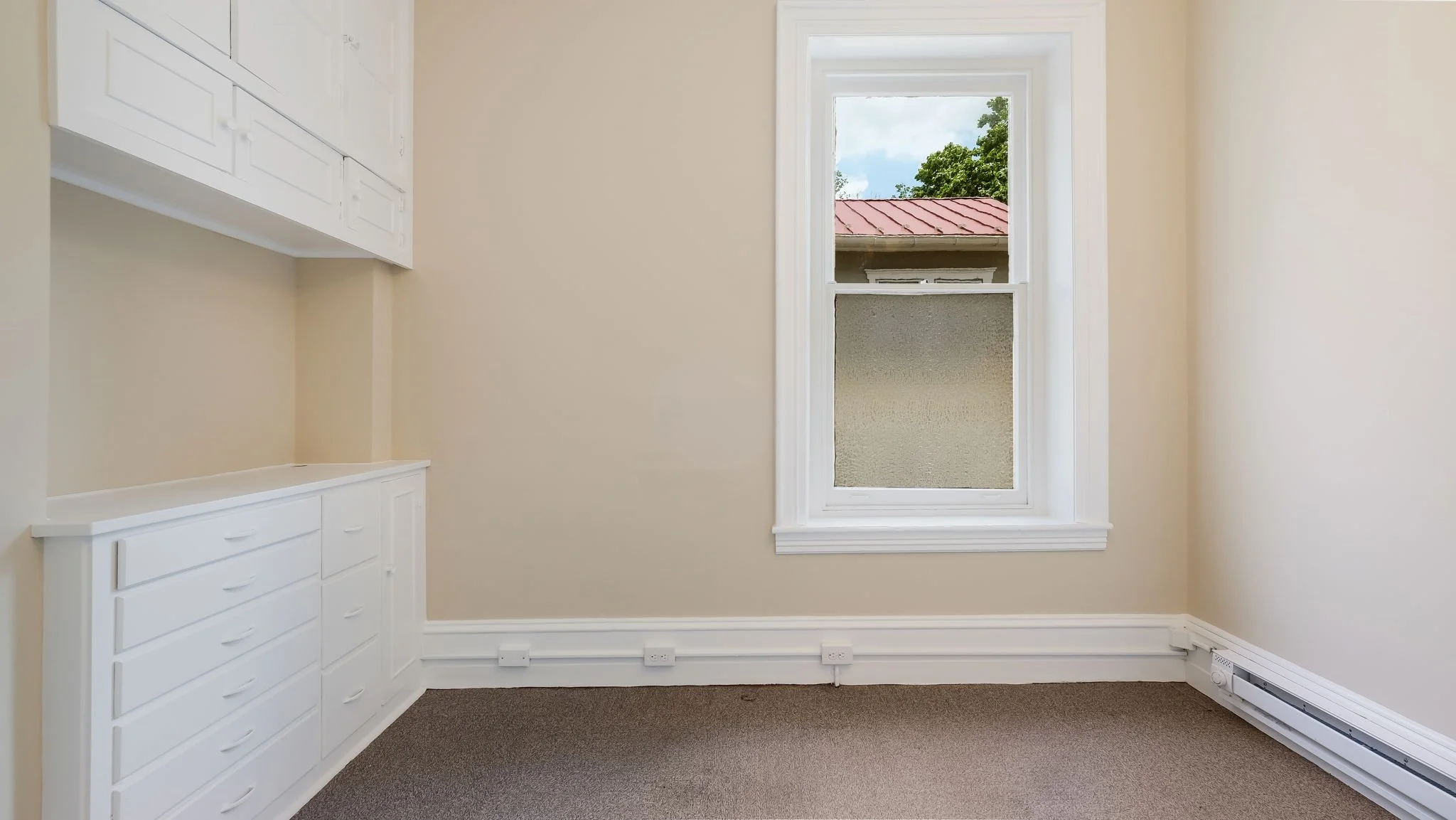 Empty room with beige walls, white built-in cabinets, a window with a view of a building with a red roof, electrical outlets along the base of the walls, and a carpeted floor.
