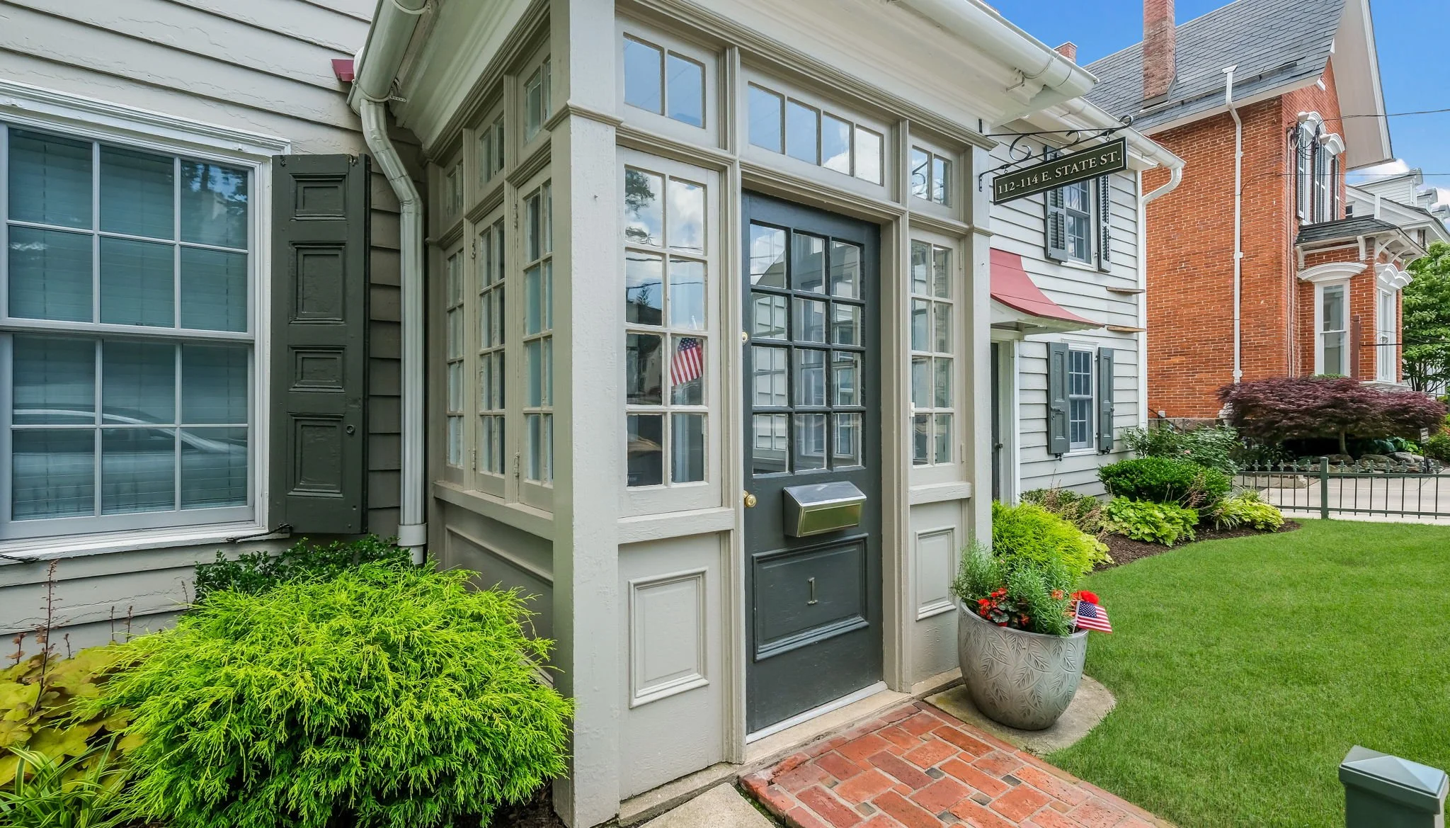 Front door of a house with a glass porch, black door with mail slot, surrounded by green bushes and flowers, with a lawn and neighboring houses.