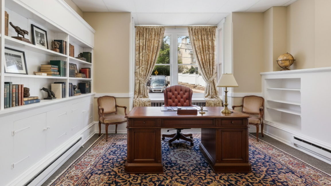 Office with wooden desk, vintage chairs, bookshelf, window with curtains, and decorative items.