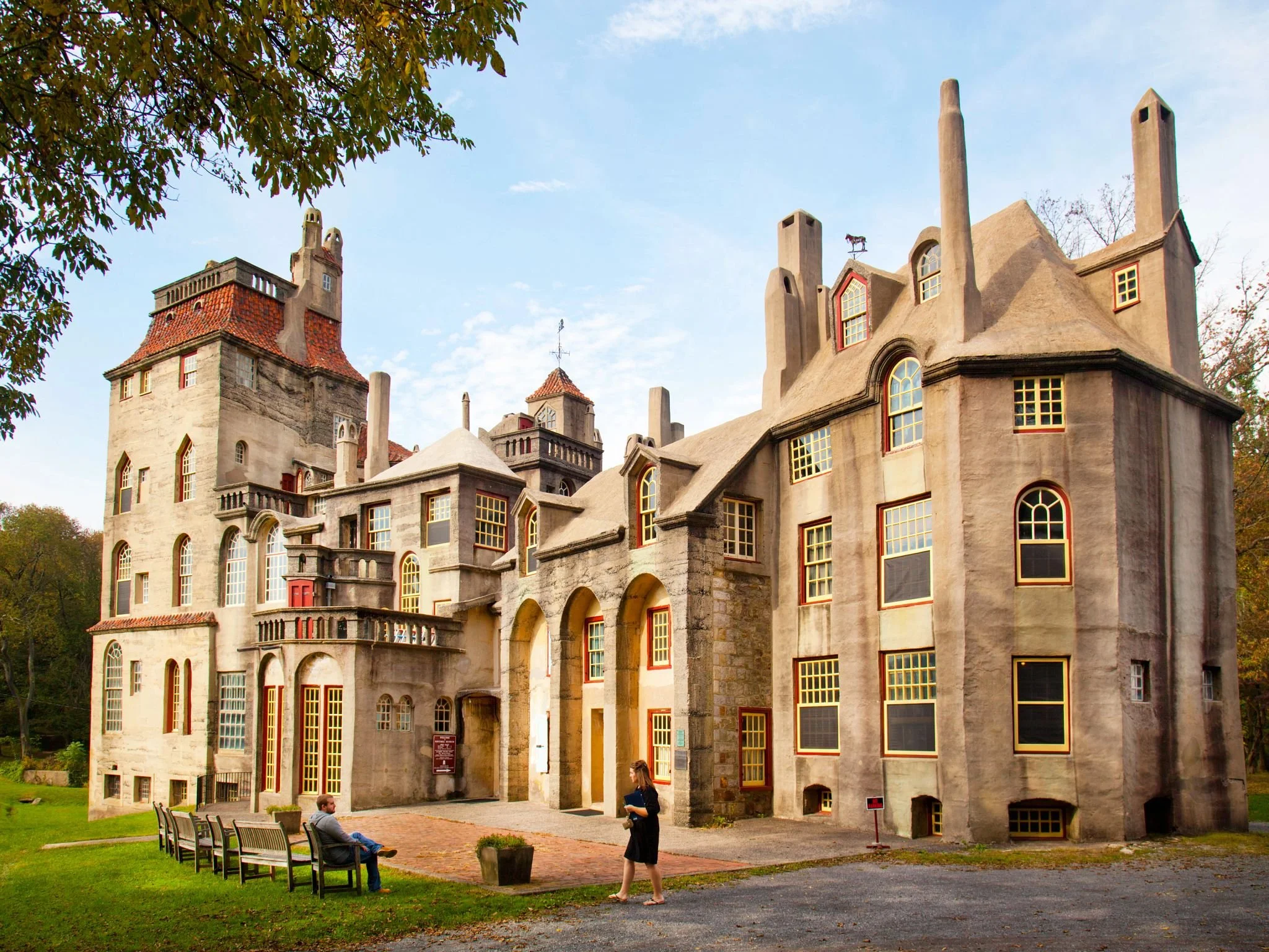 A large castle with multiple towers and windows, surrounded by trees and grass, with two people sitting on benches and one person walking outside.
