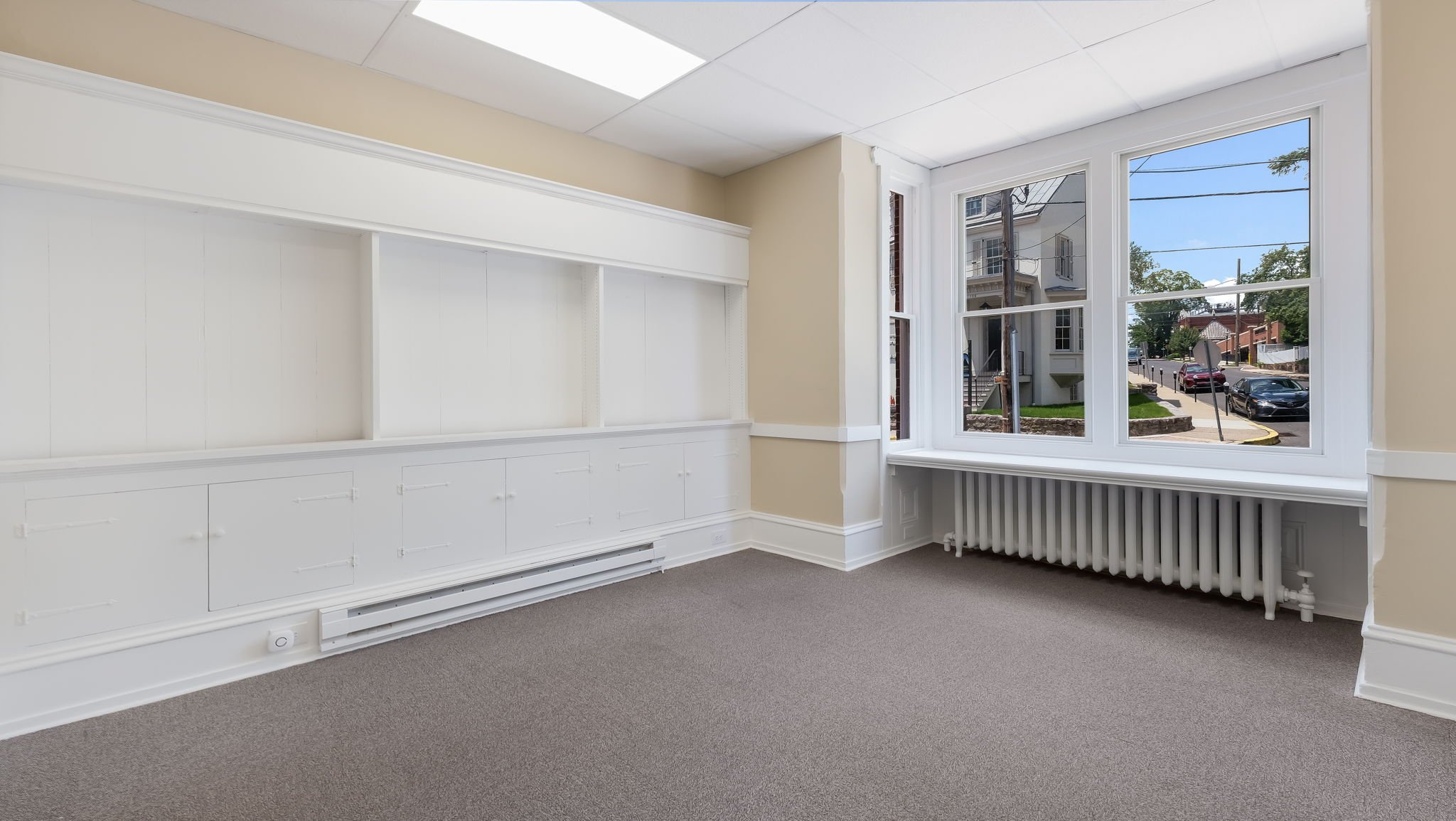 Empty room with beige walls, large window showing street view, carpeting, built-in white shelving, and radiator.