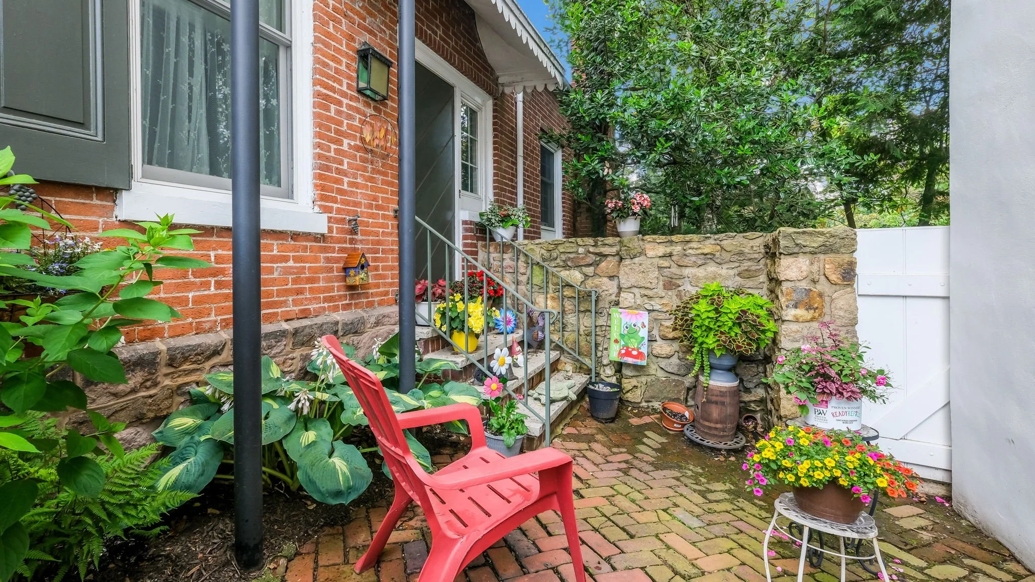 A brick patio with a red plastic chair, potted flowers, and plants near a house entrance. Green trees in the background.