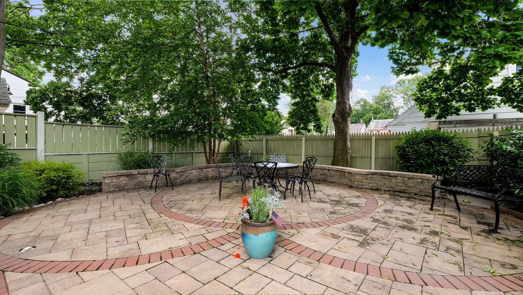 A backyard patio with a brick and stone tile surface, surrounded by green trees, shrubs, and a pale green wooden fence. There are black metal chairs and a round table, a stone low wall, and a large decorative pot with plants and a small American flag