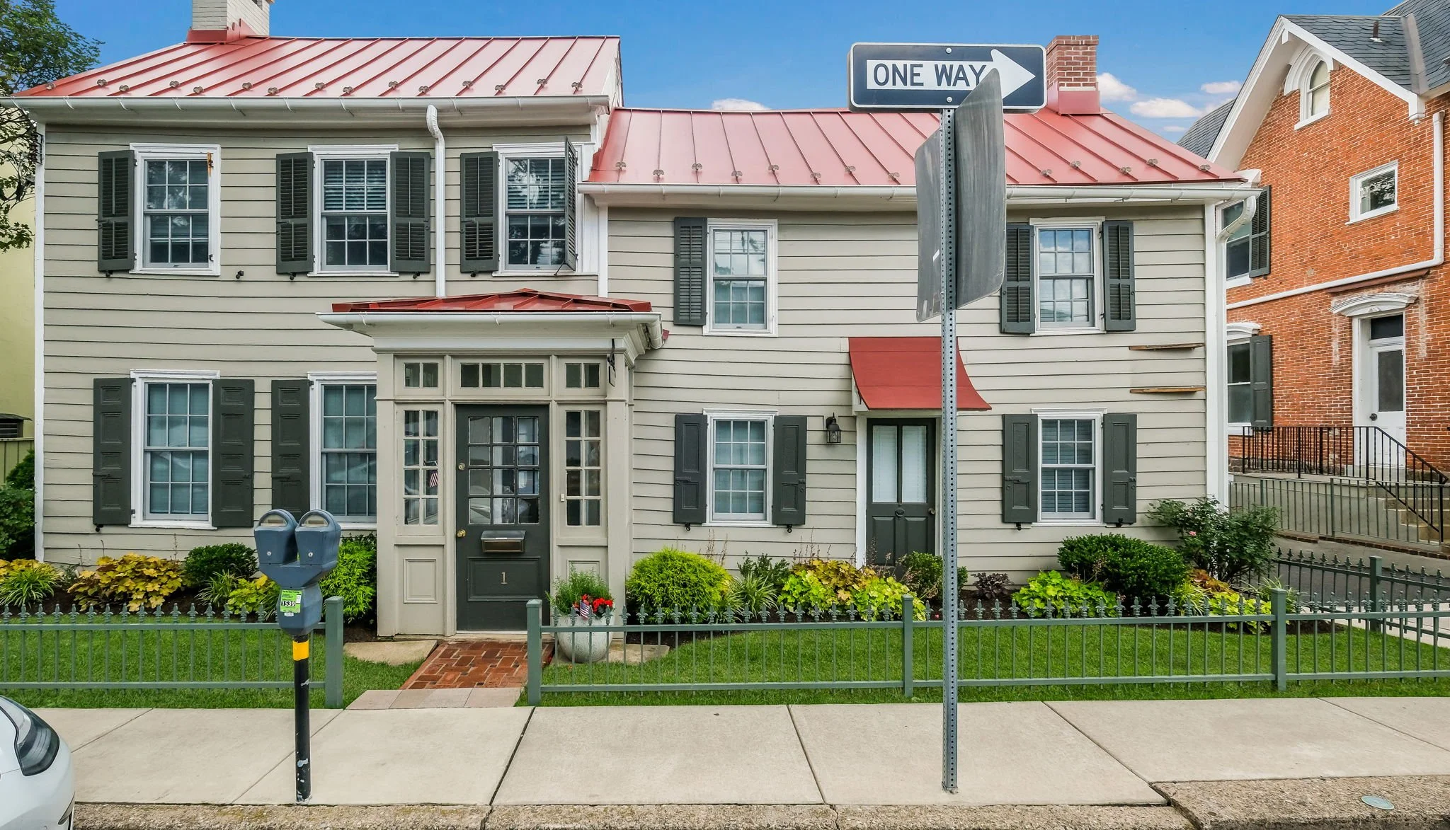 A two-story beige house with black shutters, red metal roof, and a small front garden. In front, there are two blue parking meters, a sidewalk, and a one-way street sign.