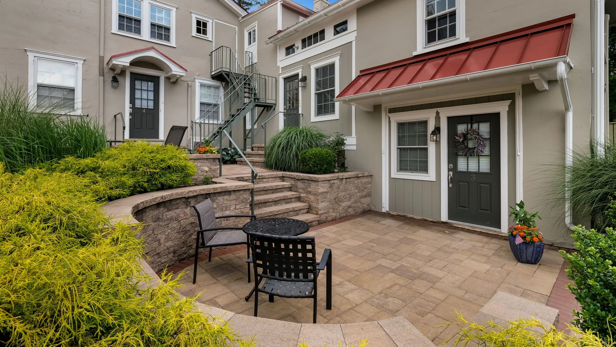 Backyard patio with black chairs, small round table, steps leading to multiple apartment doors, green plants, potted flowers, and building exterior with beige walls and white trim.