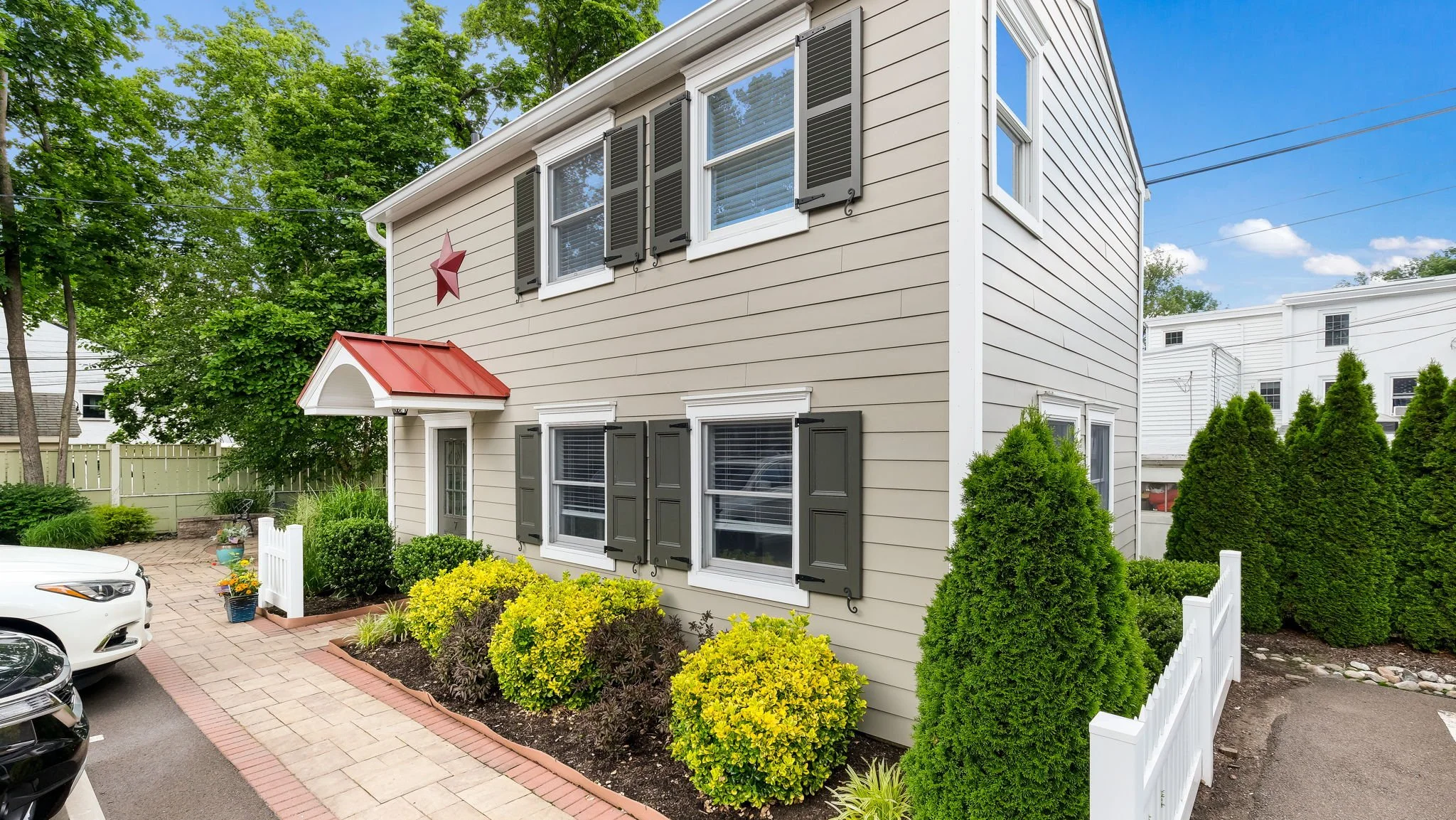 Front view of a beige two-story house with white trim, black shutters, a small red metal roof over the front door, and a red star decoration on the wall. There's a small garden with yellow and green bushes, a white picket fence, and cars parked nearb