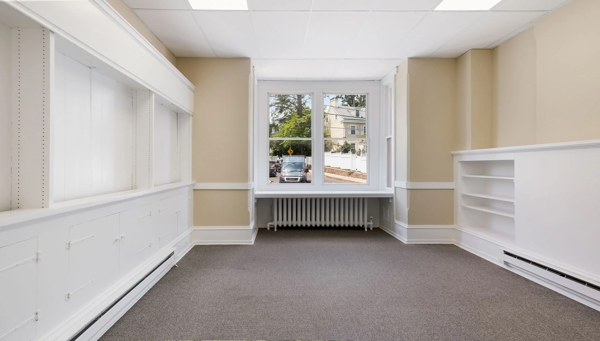Empty beige room with large window, white built-in shelves, and carpeted floor.