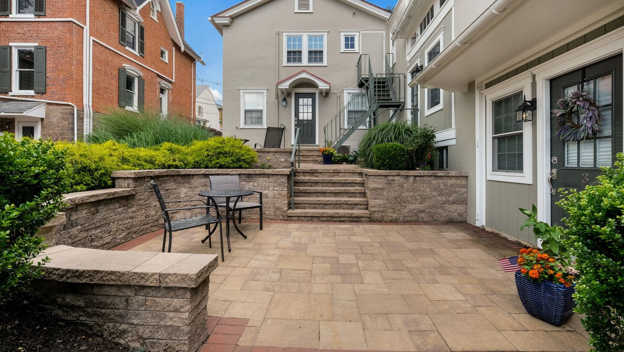 Back patio with beige stone-paved ground, a round metal table with two metal chairs, stone retaining wall with stairs leading up to a gray house with black door, green plants, and a flower box with orange flowers and American flag on the right.