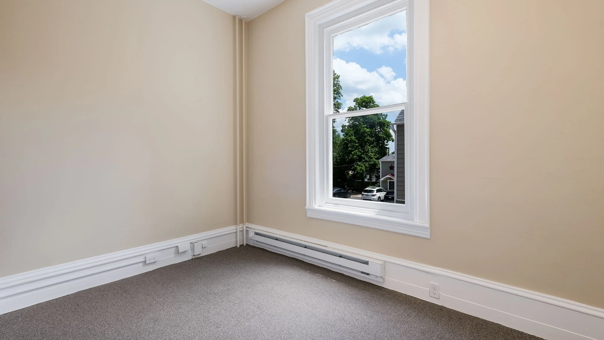Empty room with beige walls, white baseboard, window showing trees and sky, carpeted floor.