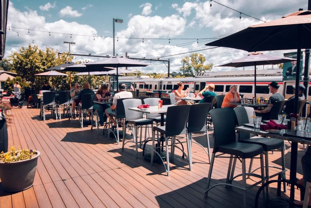An outdoor patio at a restaurant or café, with tables and chairs shaded by umbrellas. People are dining, and a train is passing by in the background under a cloudy sky.