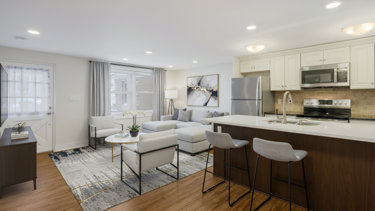 Open-concept living room and kitchen in a modern apartment with white walls, hardwood floors, a gray sofa, armchairs, a coffee table, a kitchen island with bar stools, stainless steel appliances, and large windows with curtains.