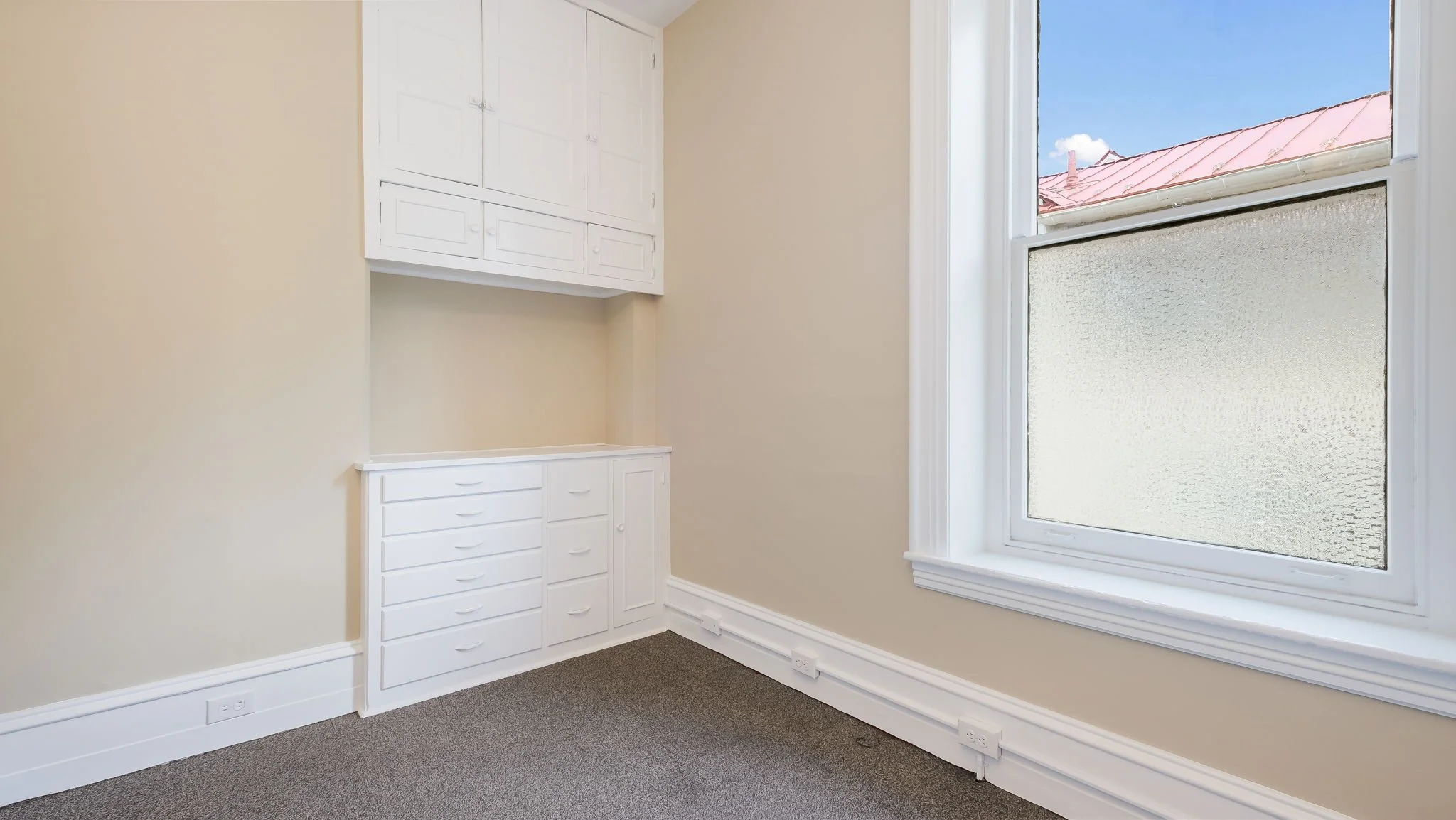 Empty room with beige walls, white built-in cabinets, a large window with semi-opaque textured glass, and carpeted floor.