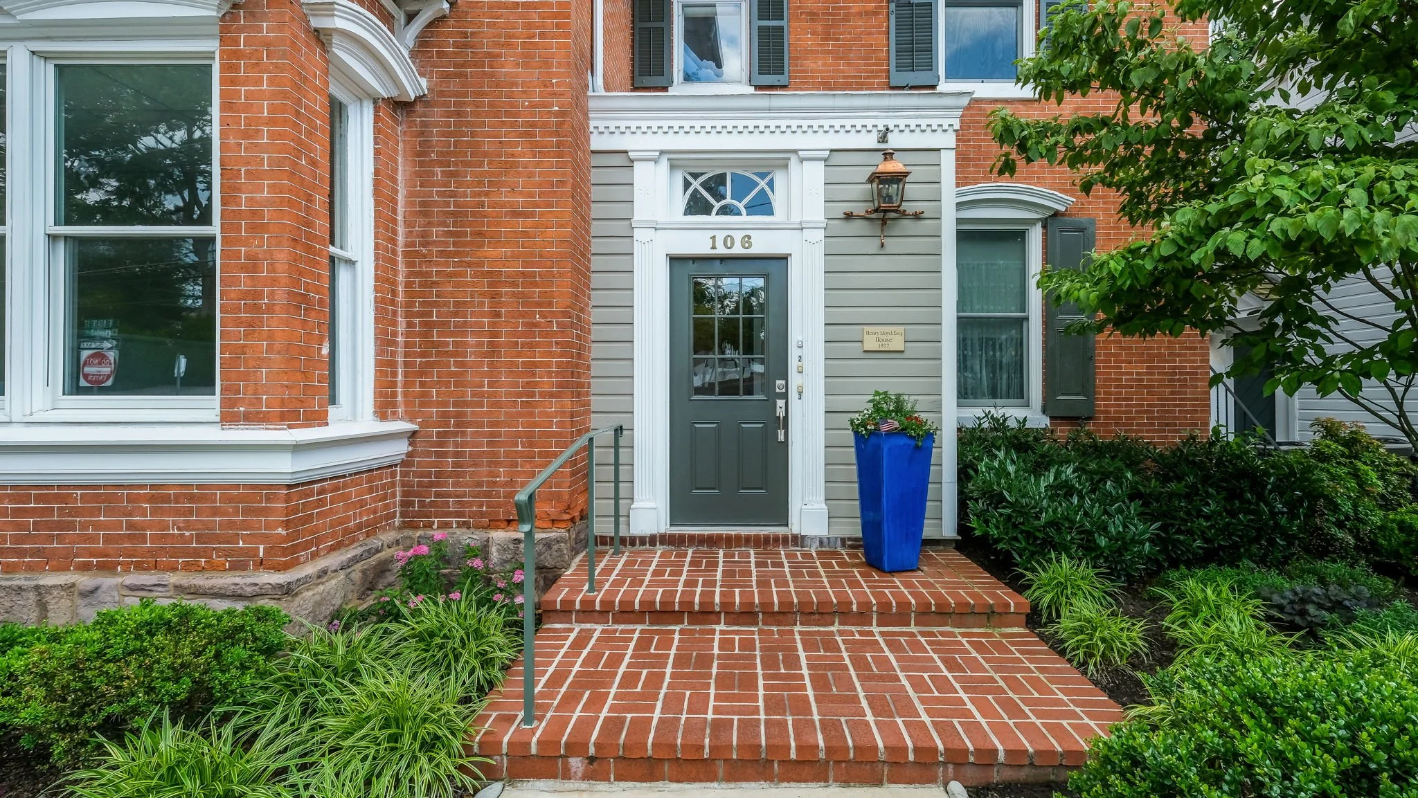 Front entrance of a house with a dark green door, brick pathway and steps, surrounded by green bushes, a blue planter with flowers, and a lantern light fixture above the door.