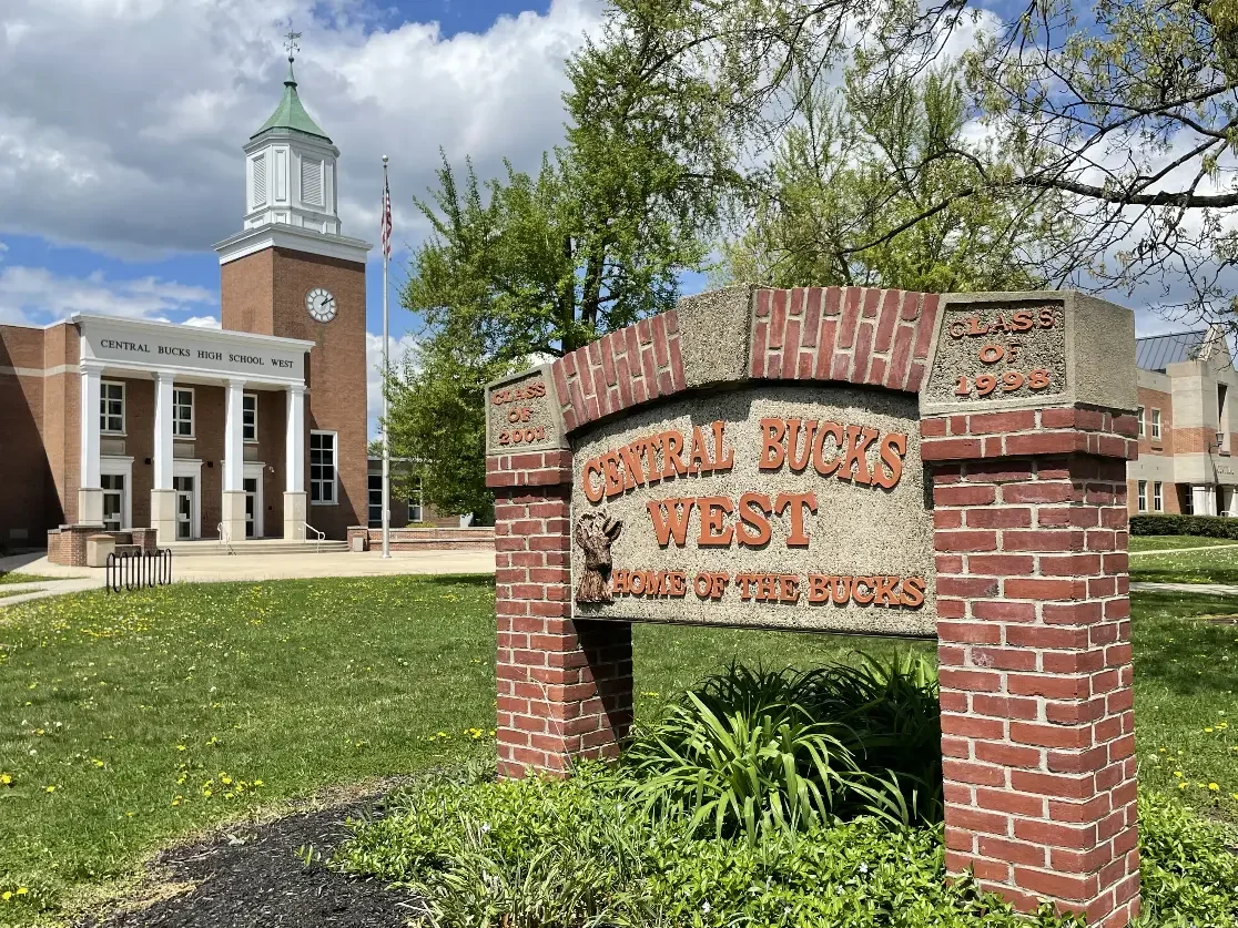 Sign for Central Bucks West High School with brick and concrete design, in front of the school building which has a clock tower, trees and a partly cloudy sky.